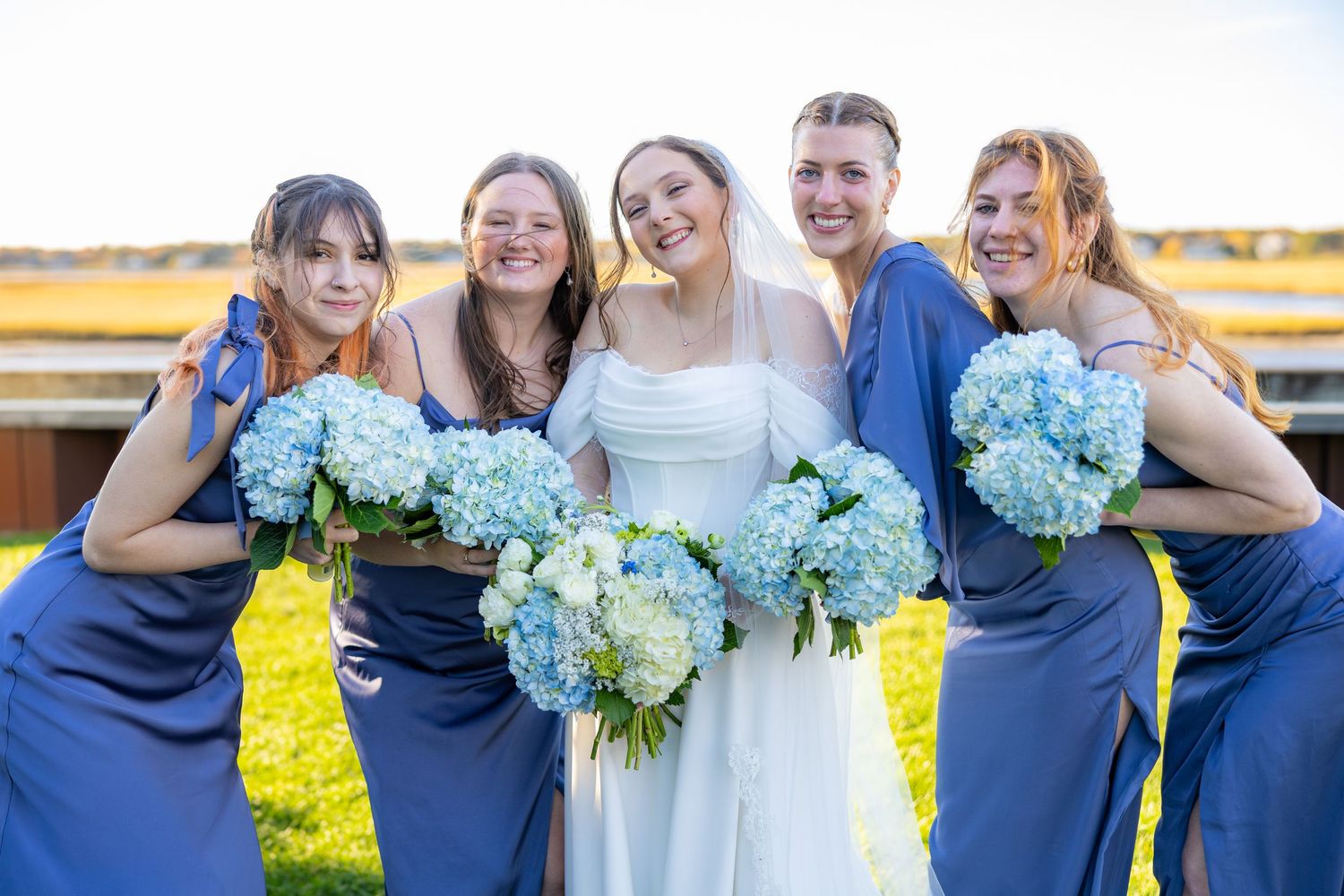 Wedding party in blue dresses poses together with light blue hydrangea bouquets in sunny outdoor setting.