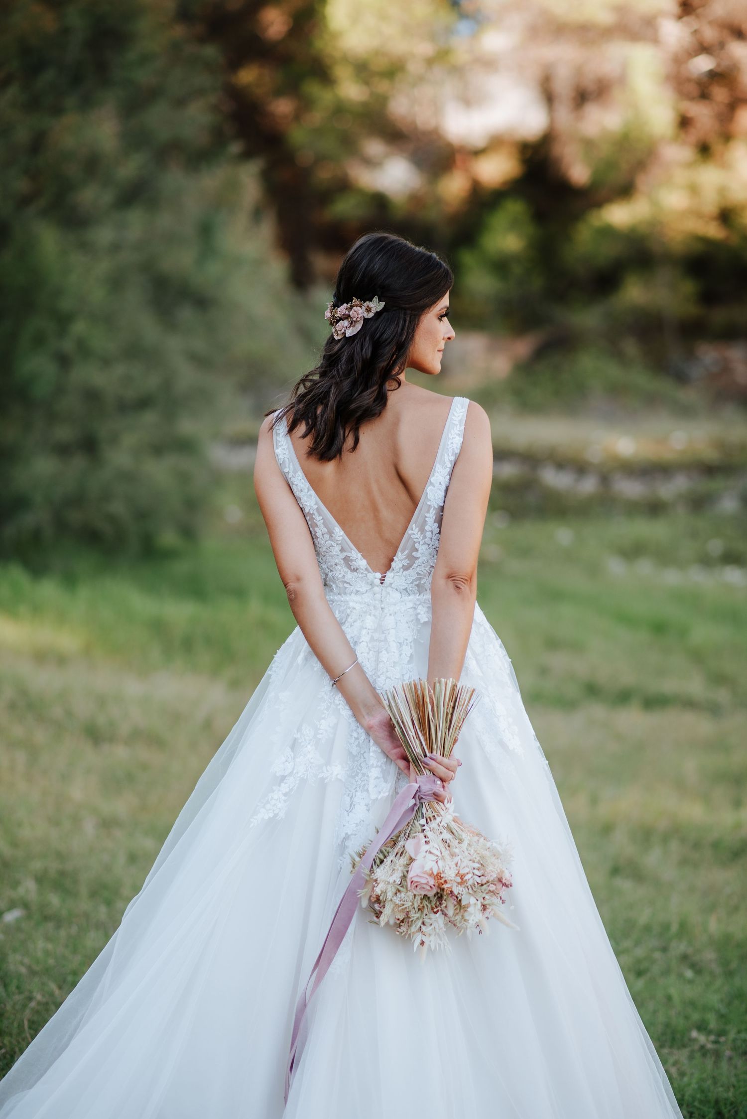 Novia de espaldas con ramo silvestre, en un campo verde. Vestido con escote en V y detalle floral. Fotografía natural al aire libre