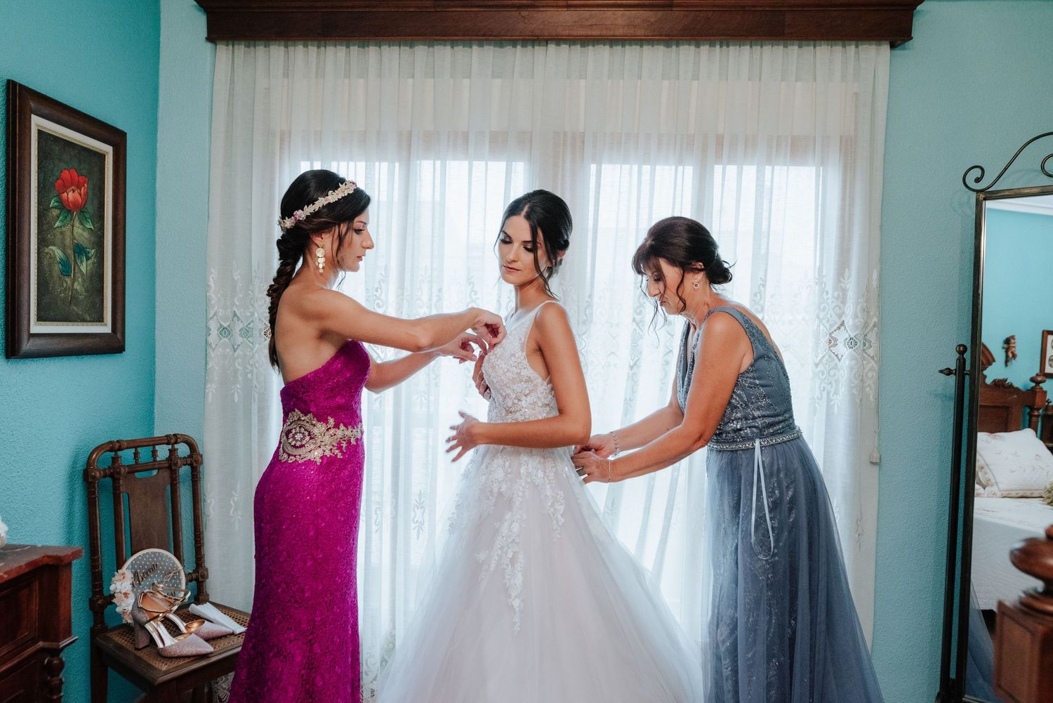 Madre y hermana junto a la ventana ayudan a la novia con el vestido