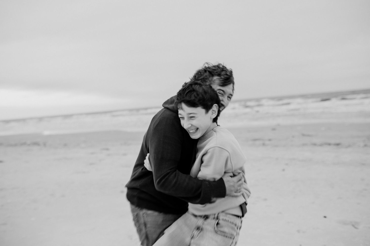 Two people share an intimate embrace on a windswept beach in black and white.