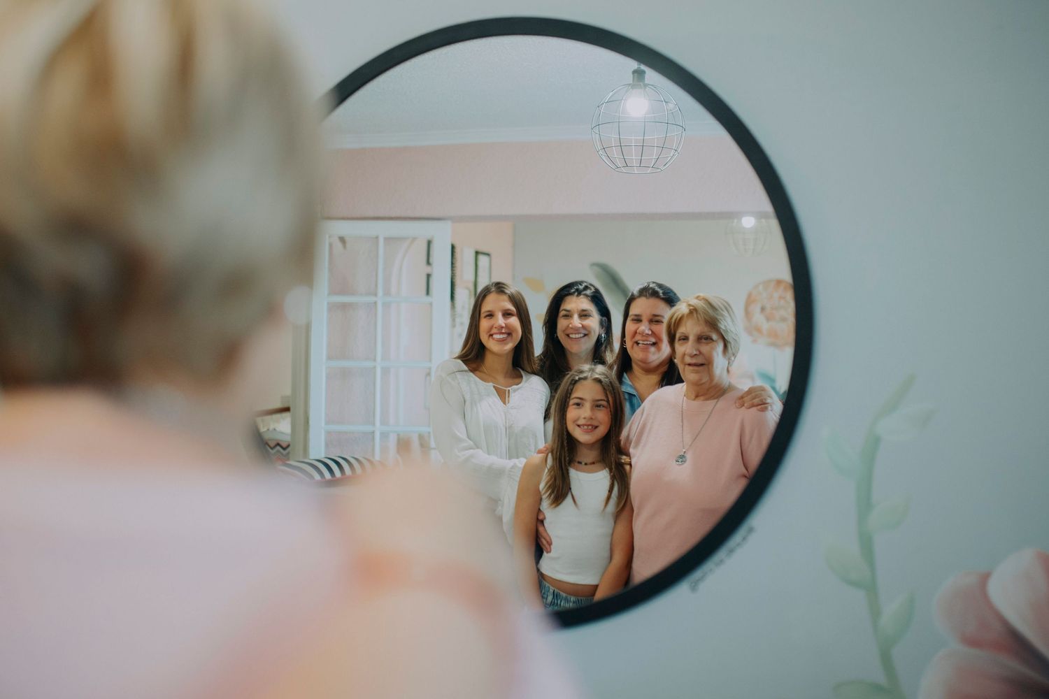 Group of women and a girl smiling together, reflected in a round mirror during a wedding preparation.