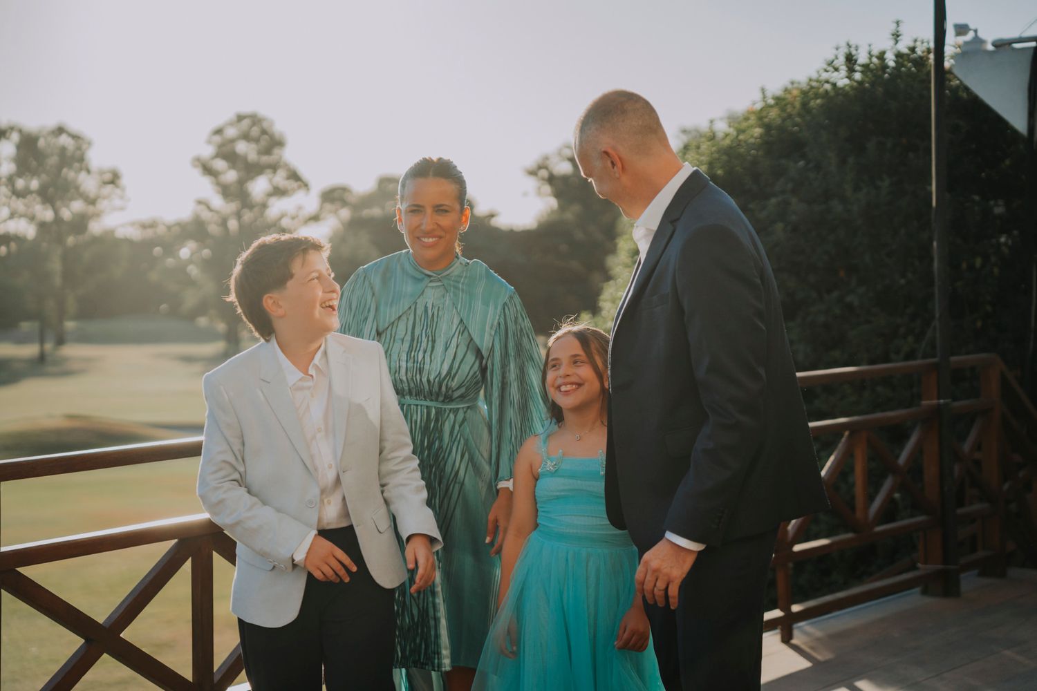 Family of four smiling and conversing outdoors at golden hour, dressed in formal attire on a wooden deck.