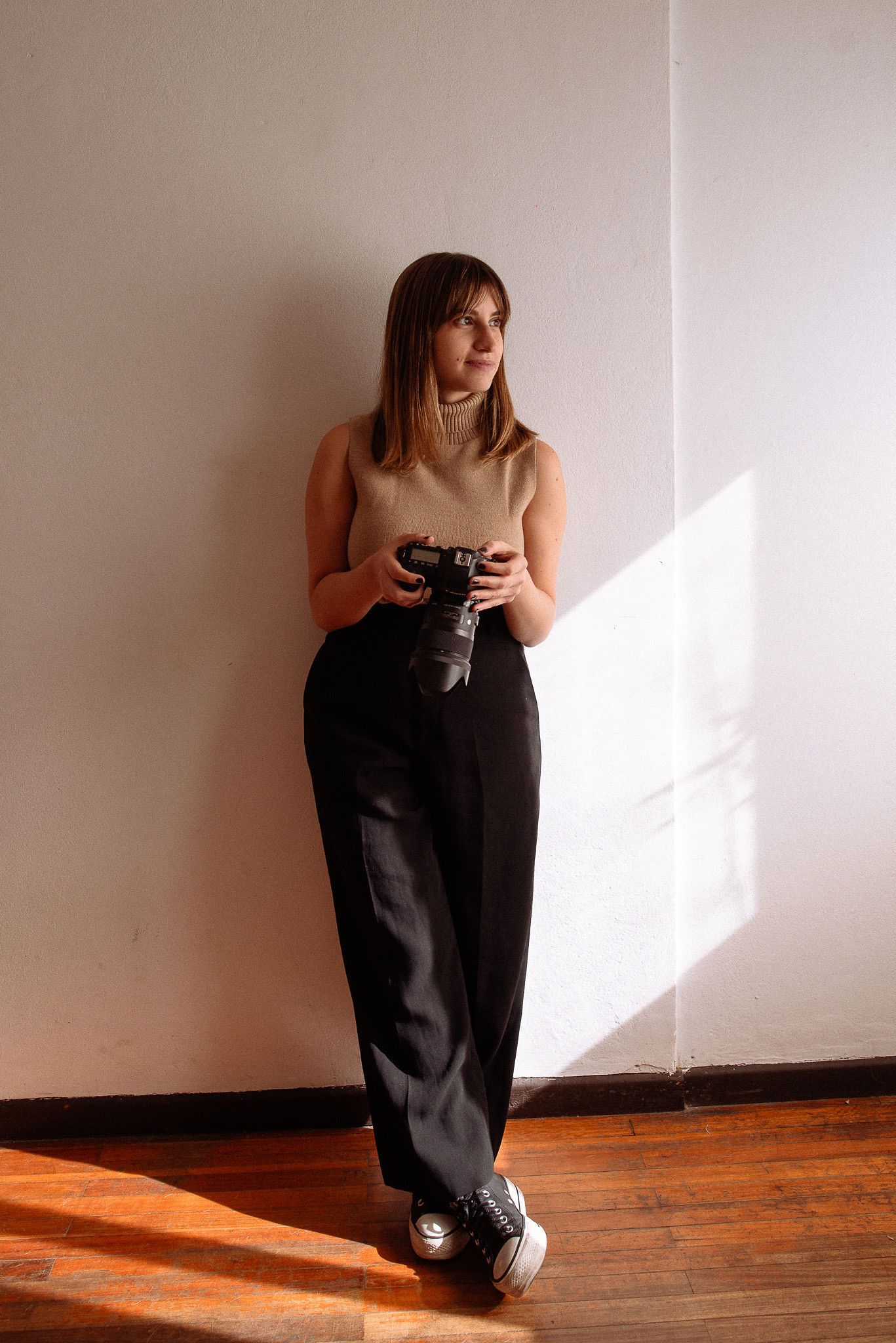 A photographer in black pants holds a camera while standing in dramatic window light.