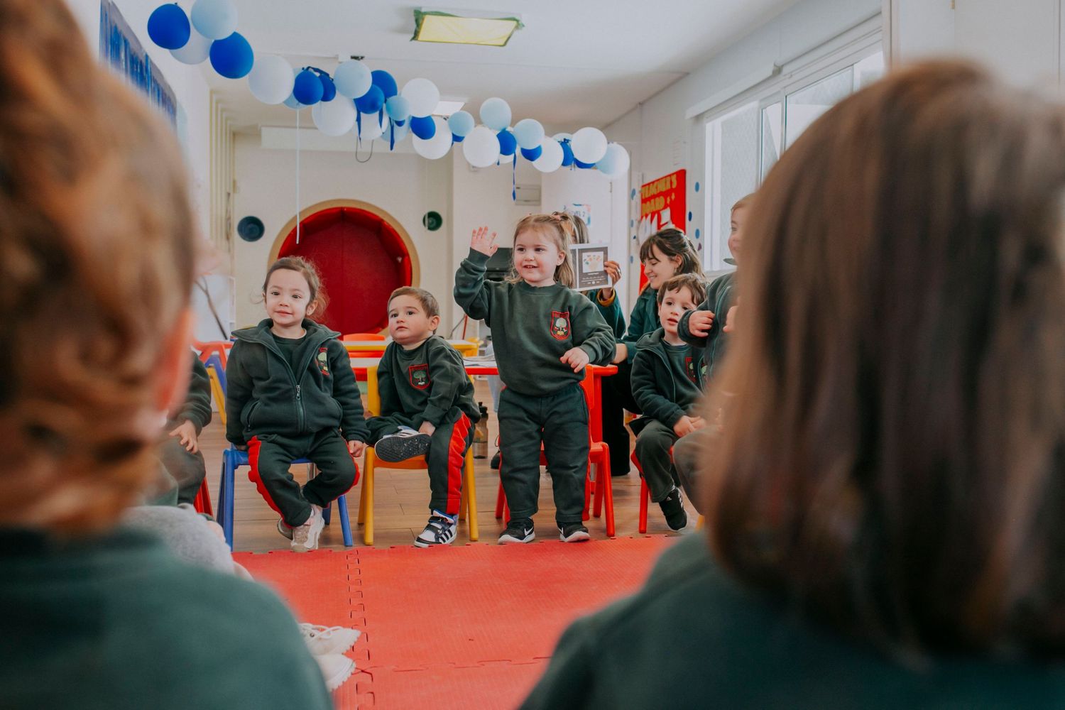 Young students in green uniforms sit in a circle on a red floor during an indoor class activity.