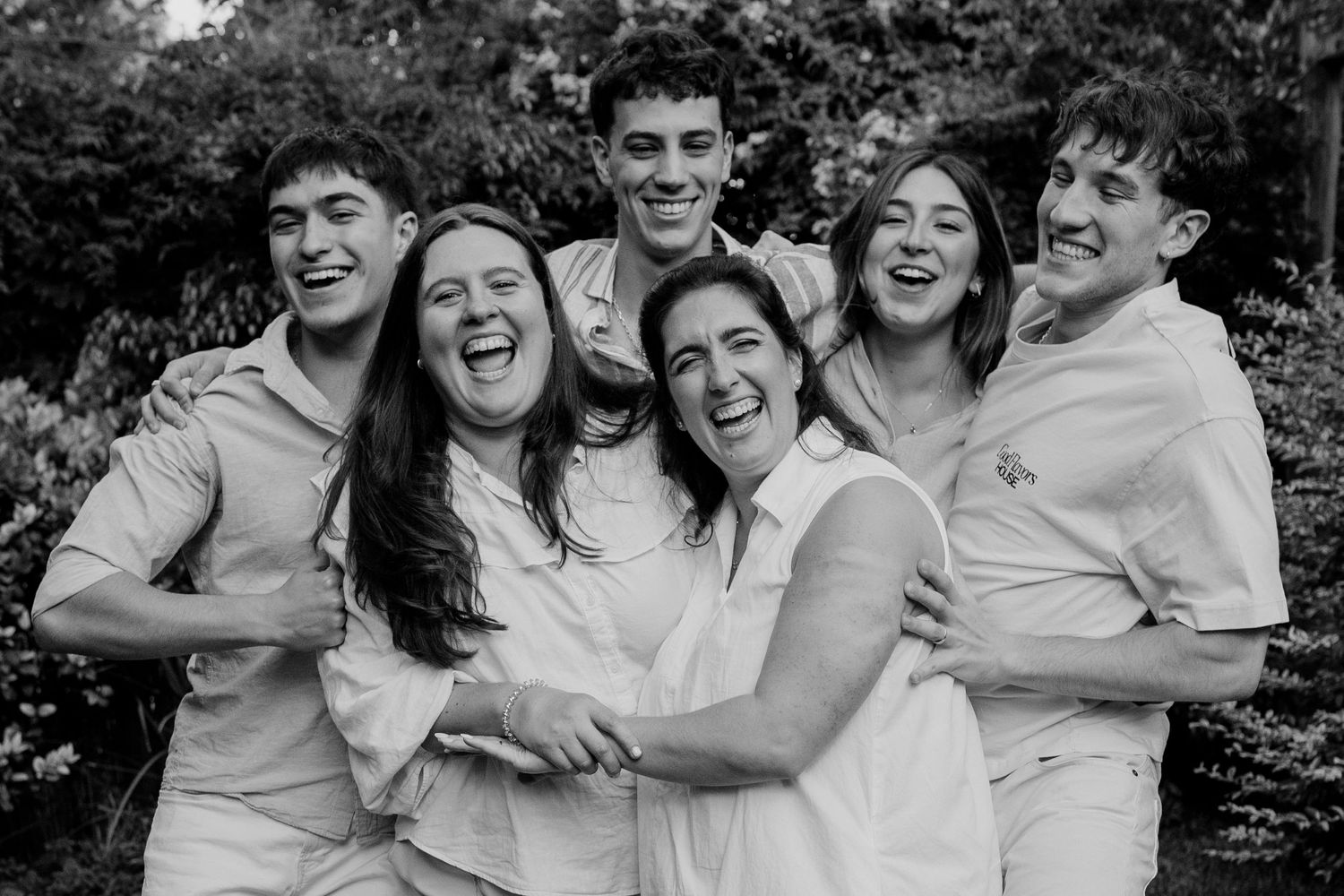 A group in white clothing laughs and embraces while posing together for a casual black and white photo outdoors.