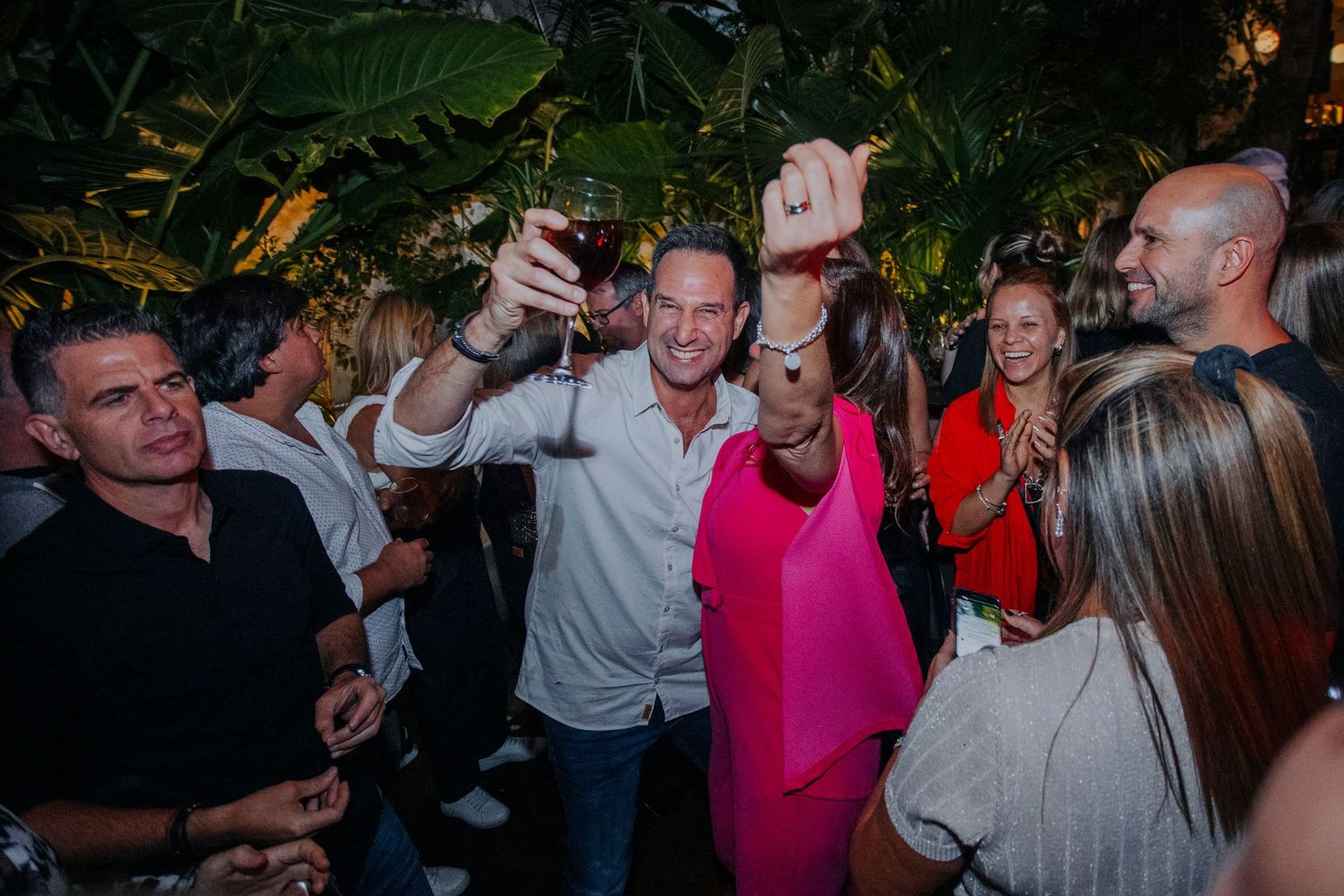 Man in white shirt raises wine glass for selfie at lively outdoor party with guests in tropical setting.