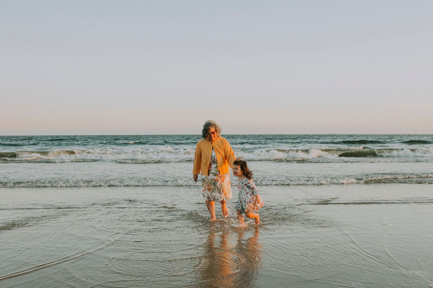Two figures wade in shallow ocean water at sunset wearing casual beachwear.