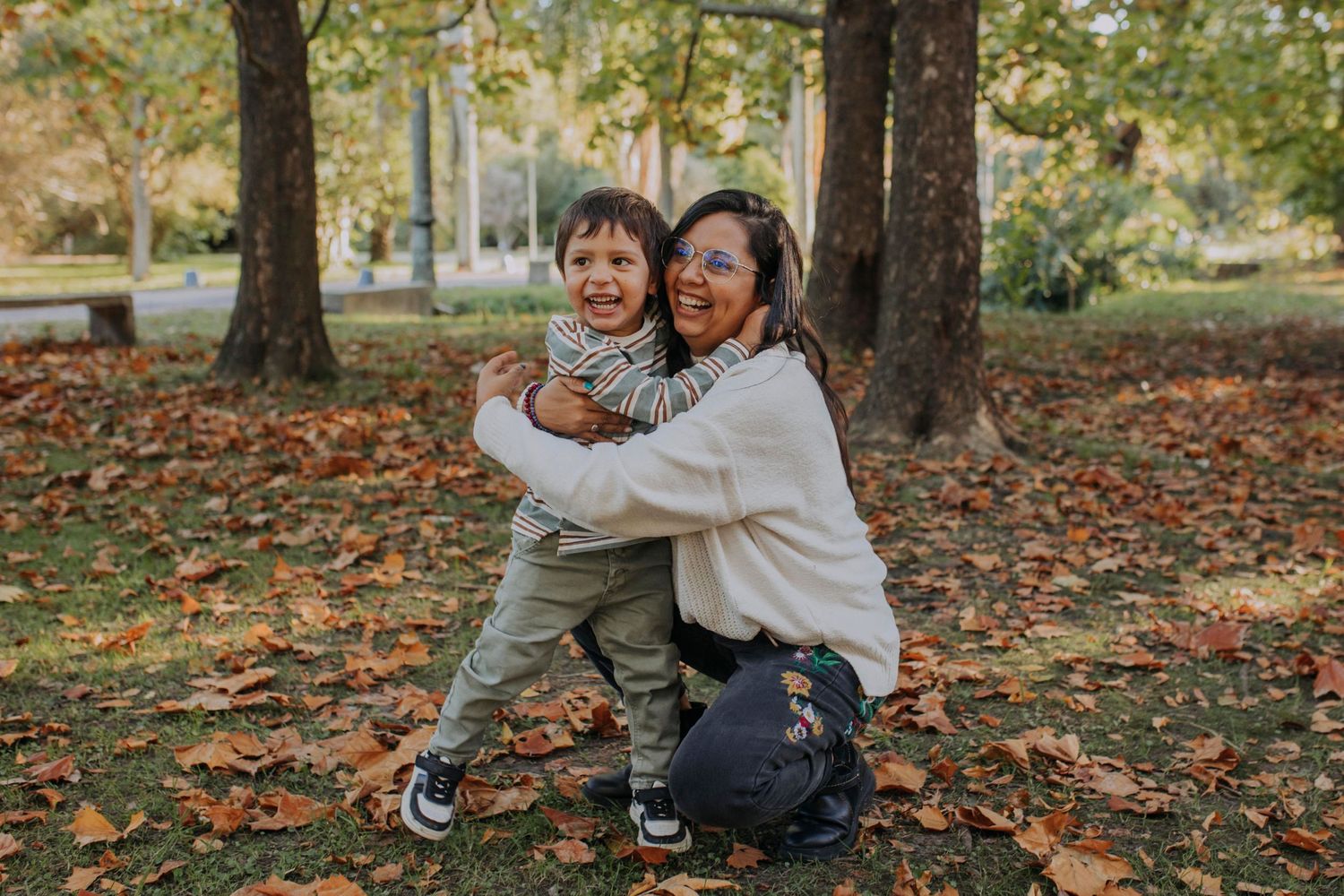 Two joyful figures share warm embrace while kneeling on autumn leaves in park setting.