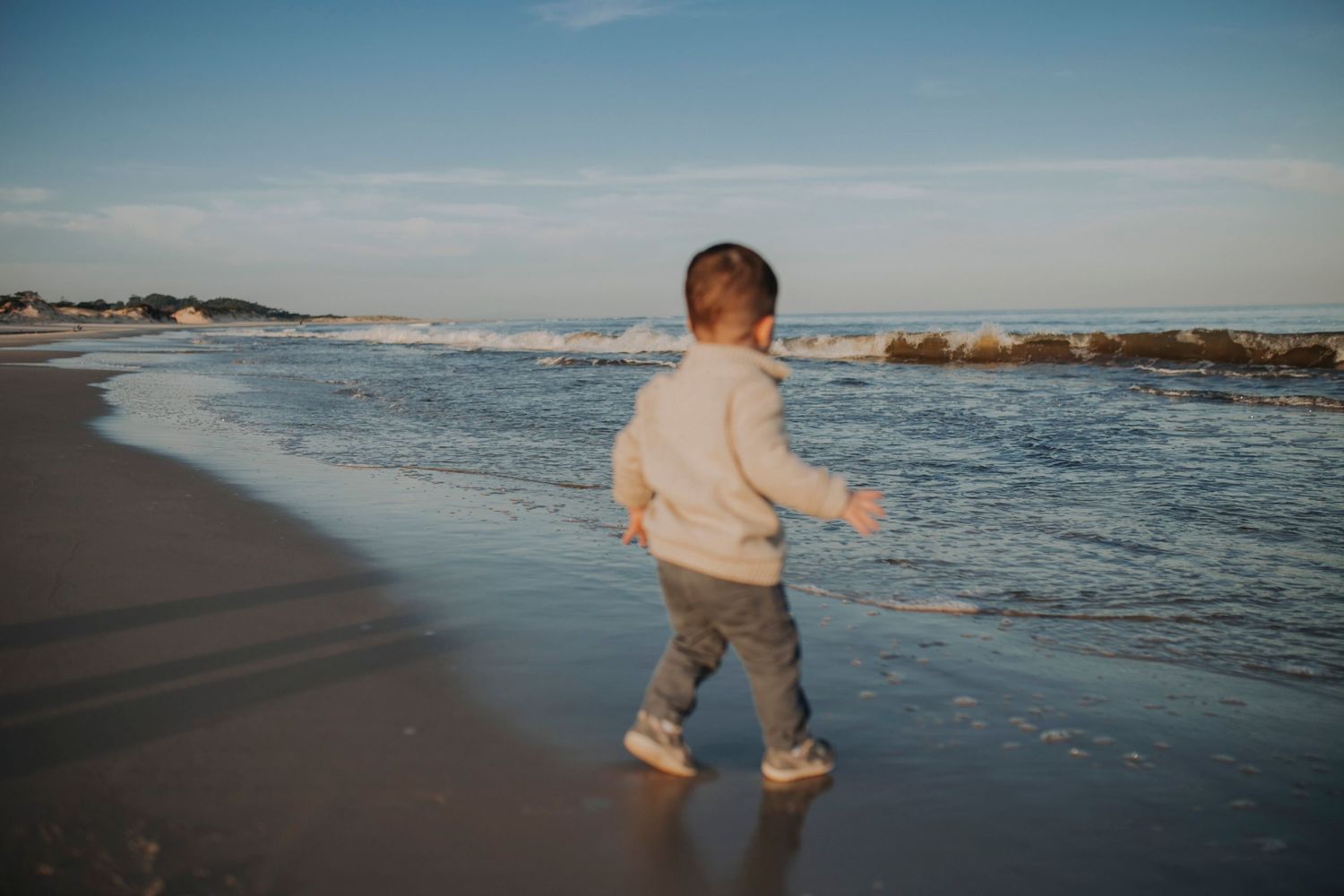 Child walking along the shoreline in a beige sweater during golden hour.