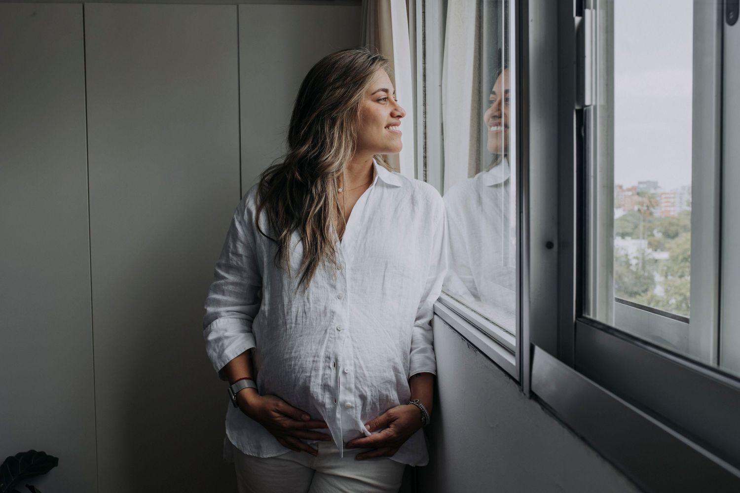 An expectant mother in white clothing gazes peacefully out a window while touching her belly.