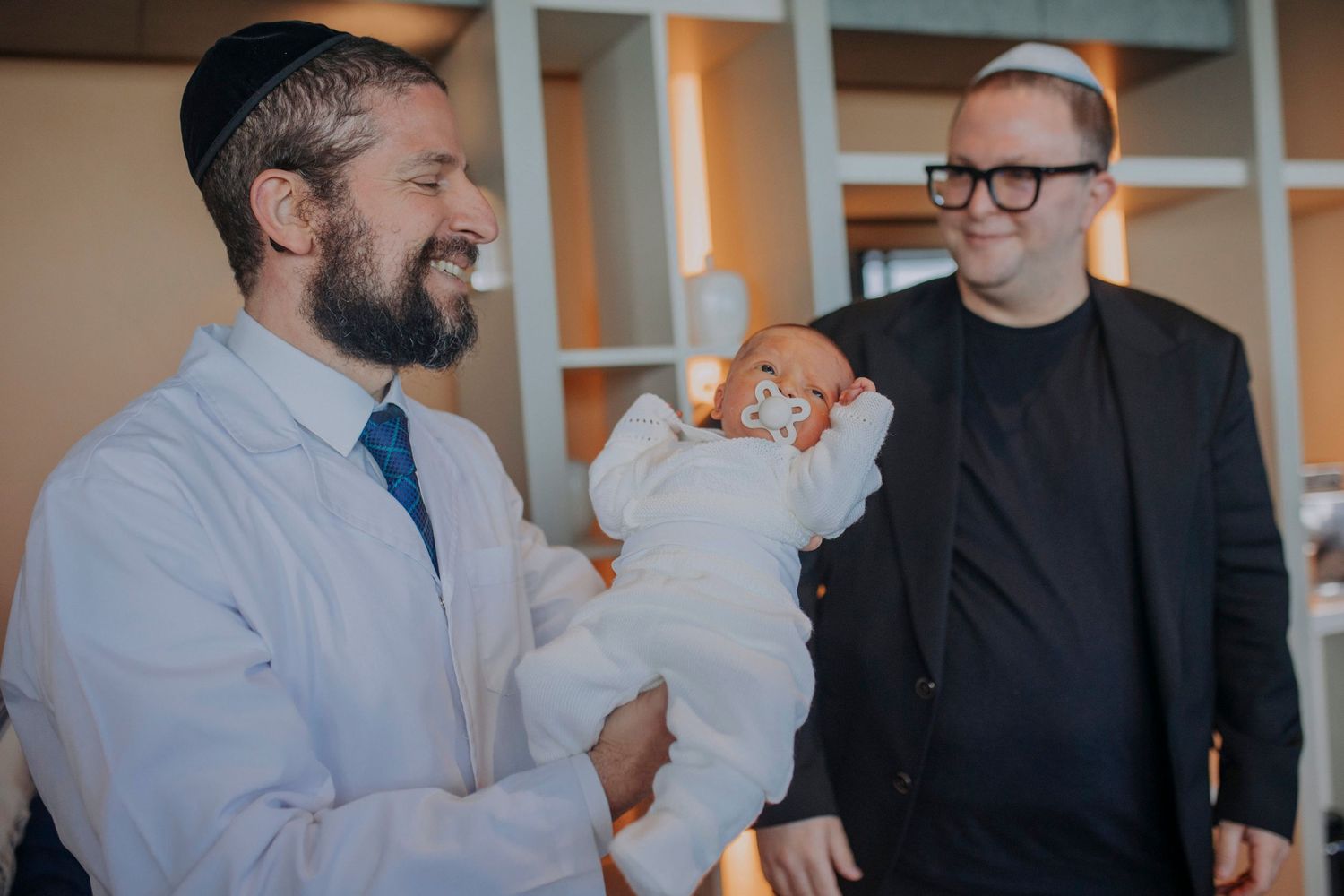 Two men smiling, one holding a newborn baby dressed in white during a Jewish ceremony indoors.