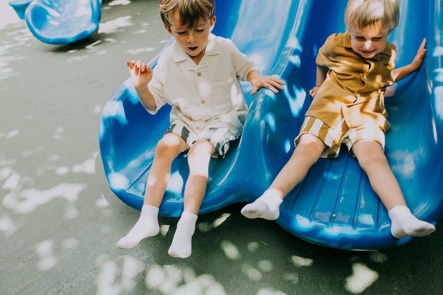 Two children in casual attire having fun on a bright blue playground slide on a sunny day.
