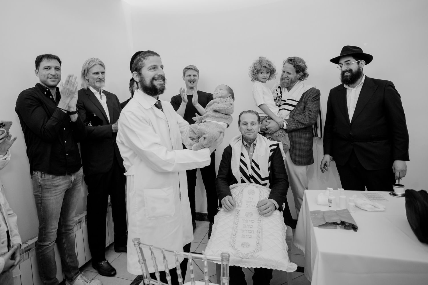 Jewish ceremony with rabbi holding infant surrounded by family members in formal attire in black and white photo.