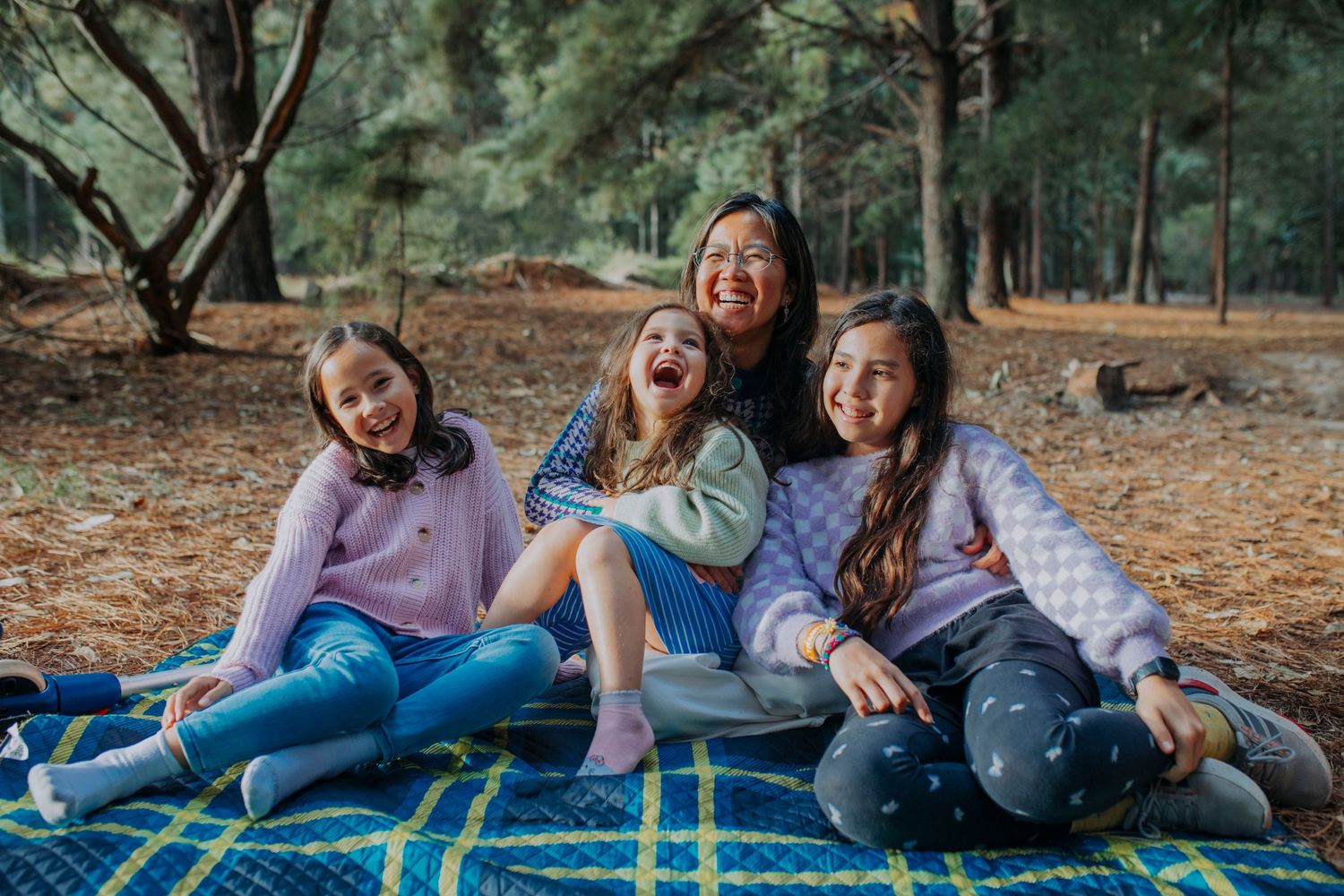 Four girls smiling and laughing together on a blue blanket in a pine forest during a fun outdoor picnic.