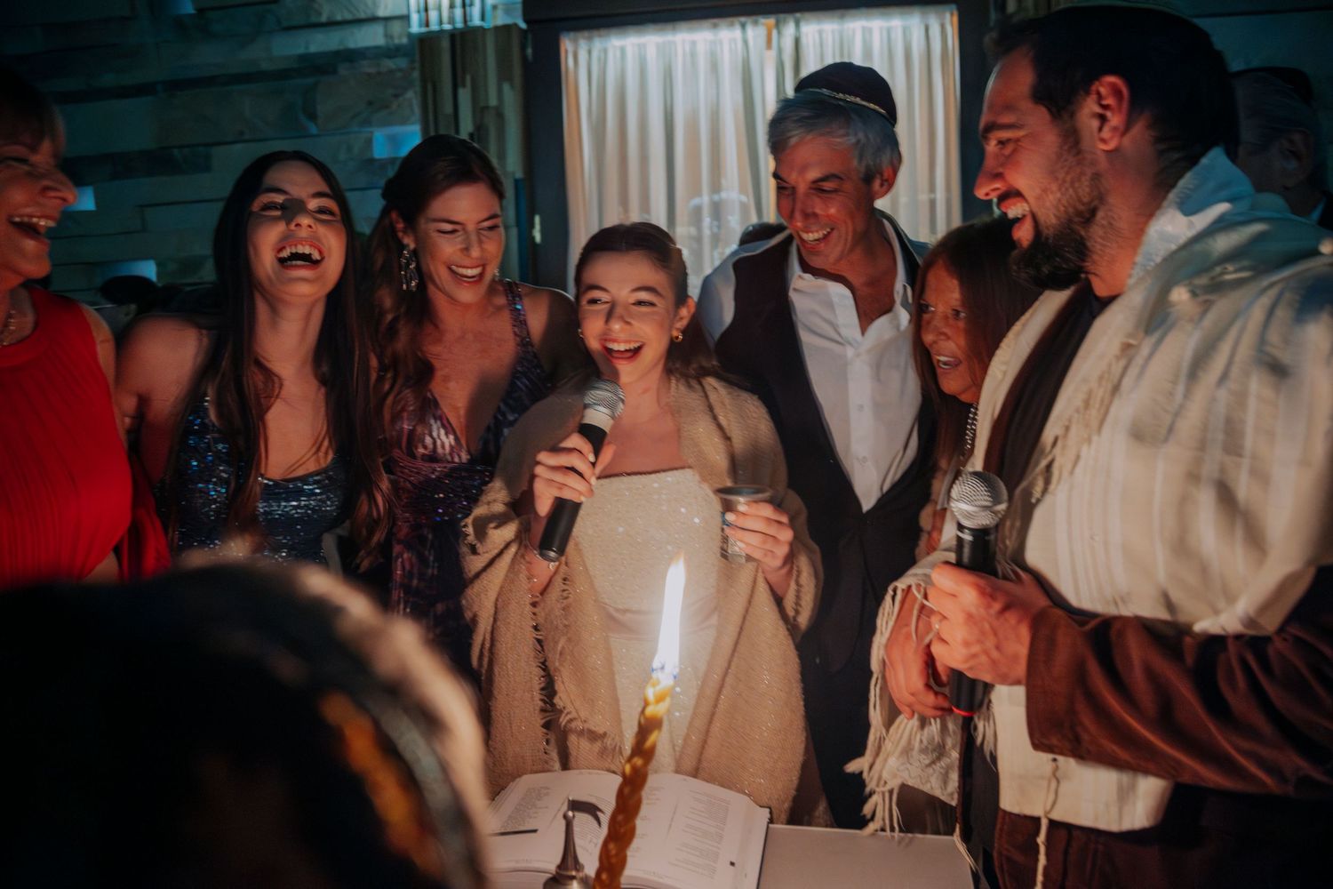 Group of joyful people celebrating at a party, with candles lit and someone holding a microphone indoors.