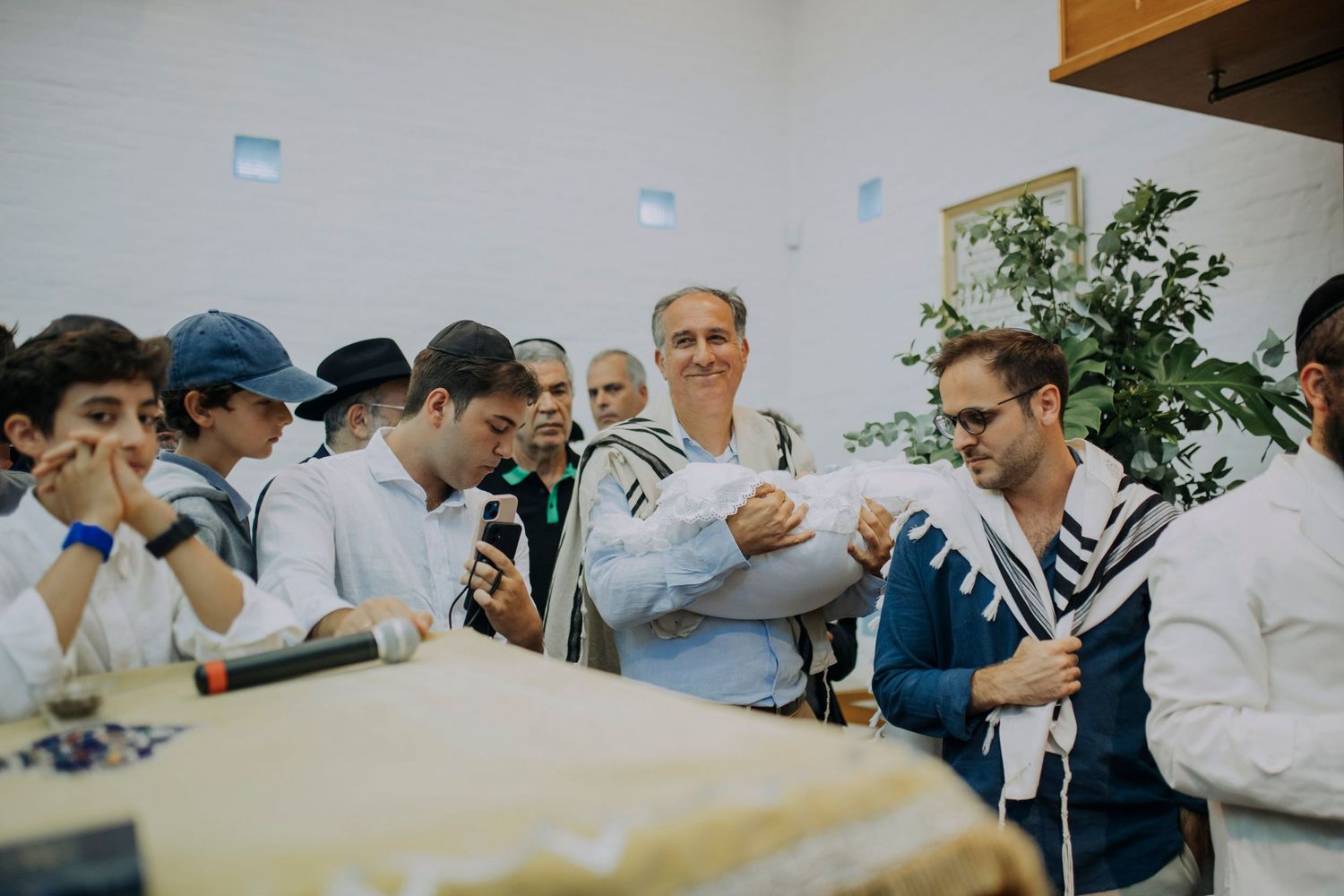 Jewish men in tallitot gather around a table during a Bar Mitzvah ceremony in a synagogue.