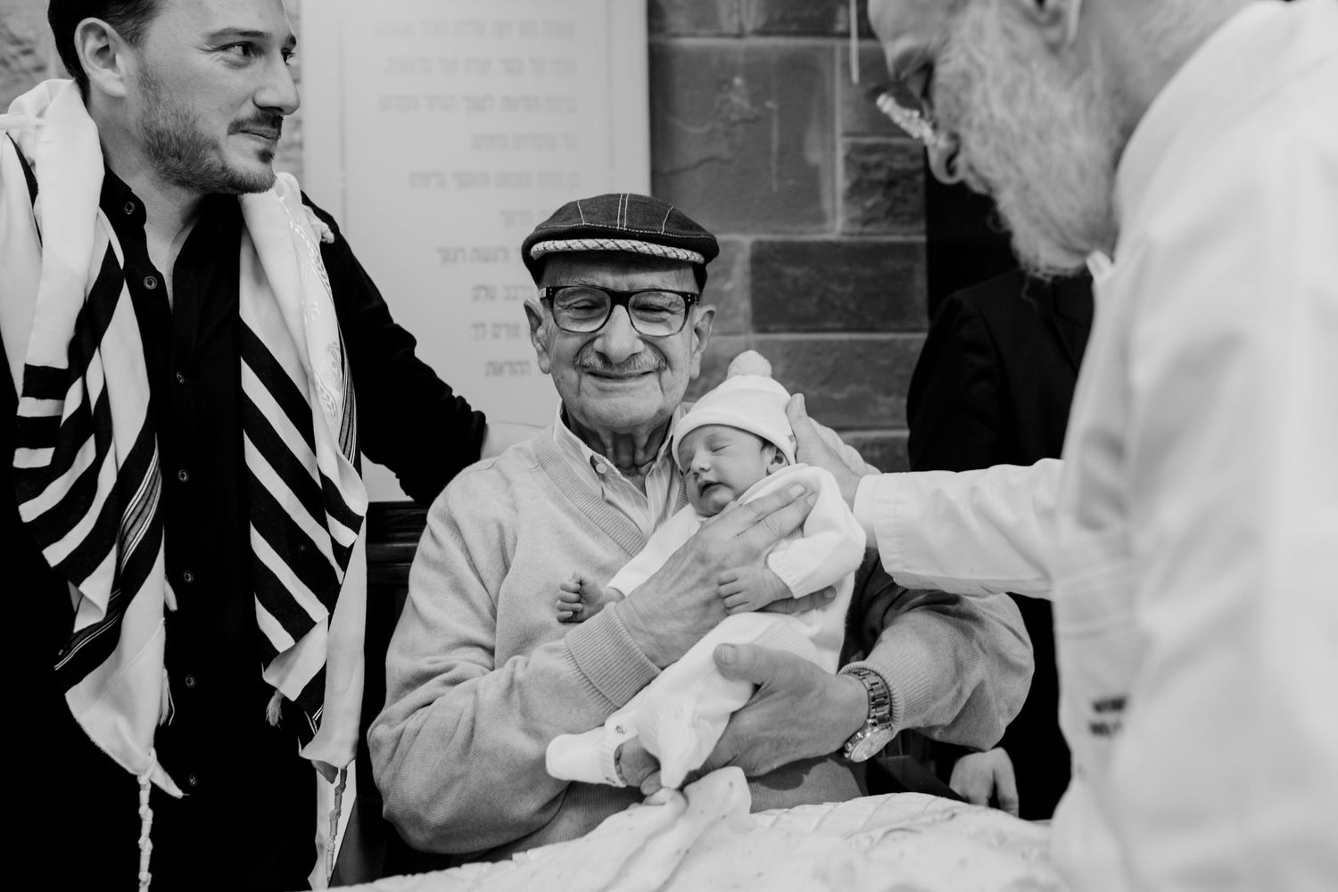 Elderly man holds newborn baby during Jewish bris ceremony, surrounded by family in prayer shawls.