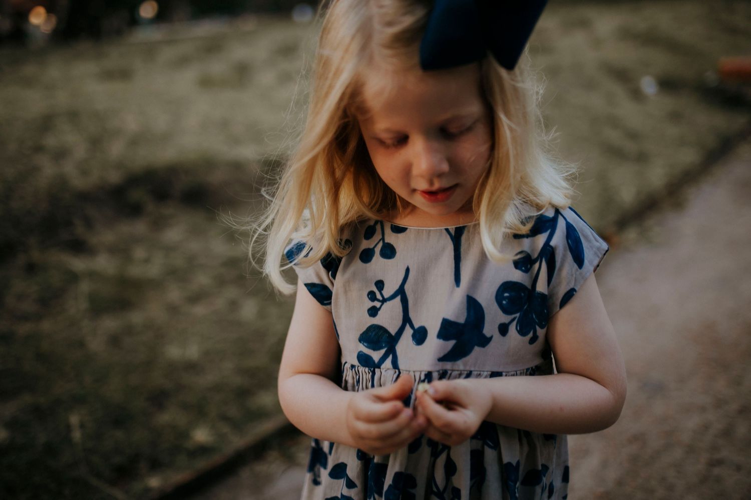 Young child in floral dress looking down thoughtfully in outdoor setting.