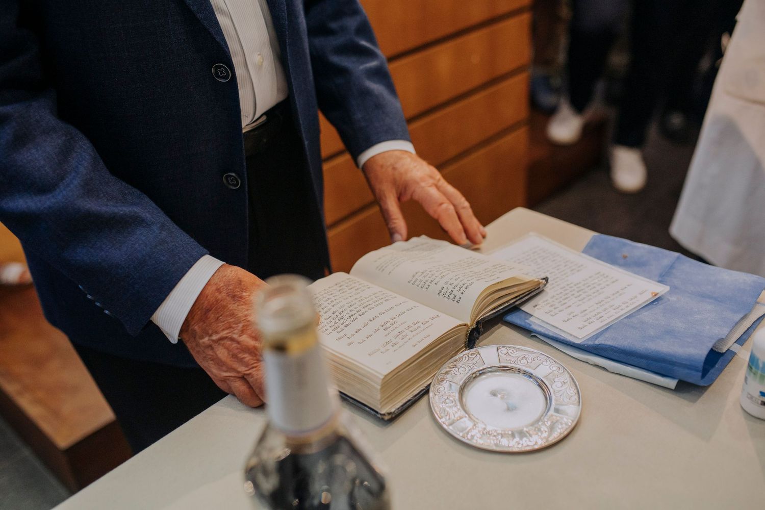 Man in blue suit reading open Bible on altar with decorative plate and candle during religious ceremony.