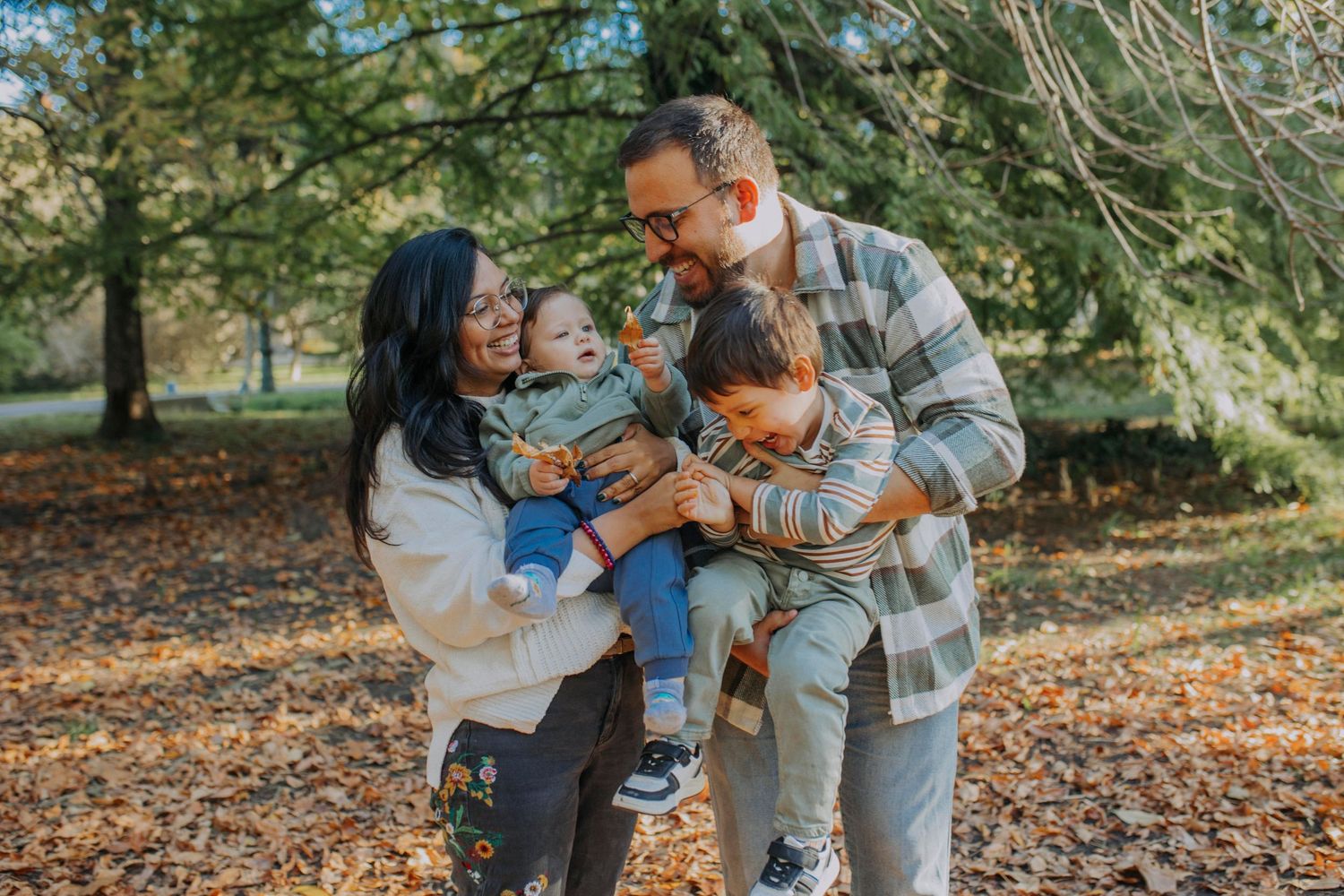 Happy family of four enjoying autumn park, parents holding two young children among golden fallen leaves and trees.