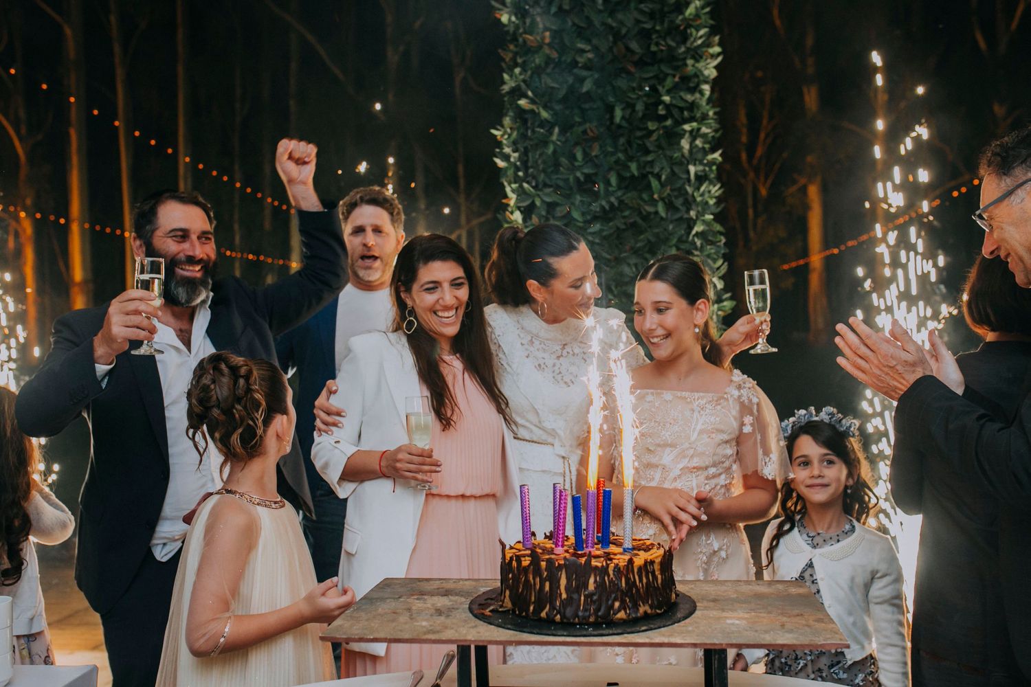 A festive celebration around a birthday cake with sparklers at an indoor evening party with string lights and trees.