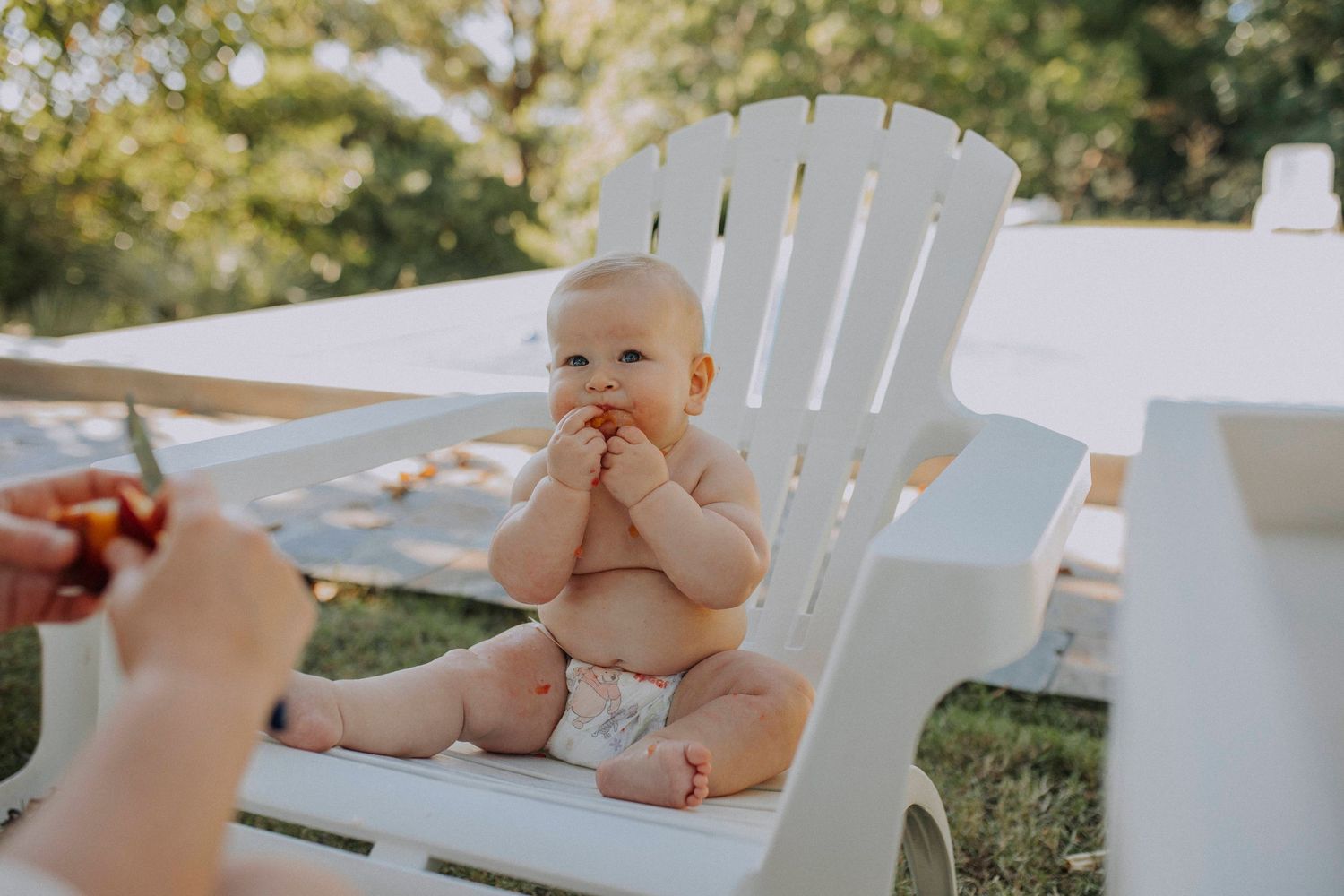 Small baby sits on white Adirondack chair in sunny backyard wearing only a diaper.