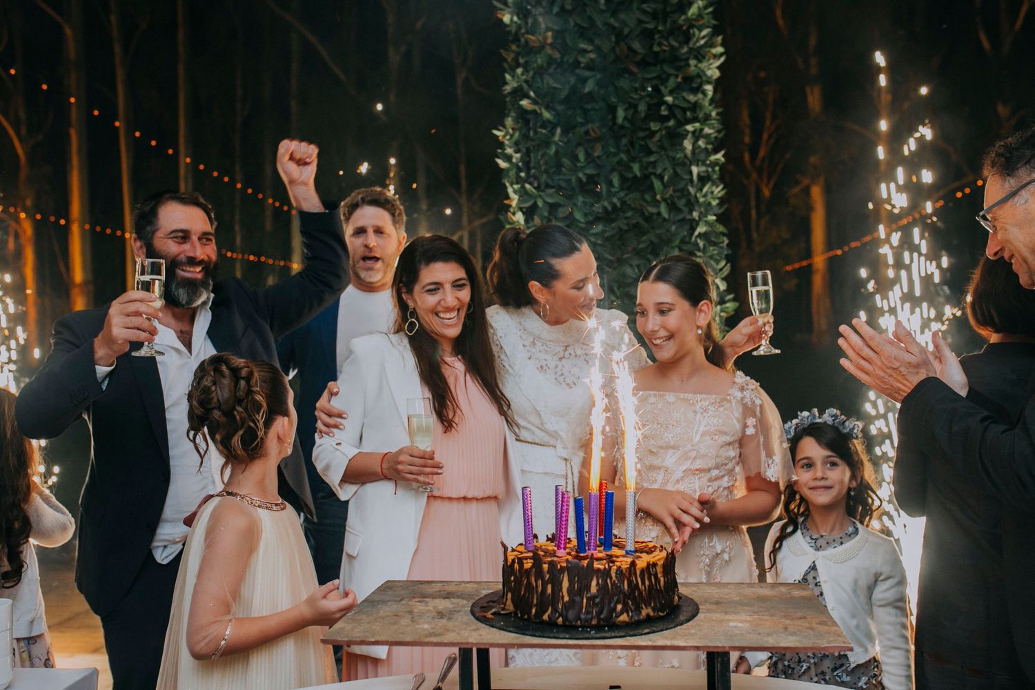 Group celebrating around a dark birthday cake with lit candles and sparklers at an elegant outdoor evening party.