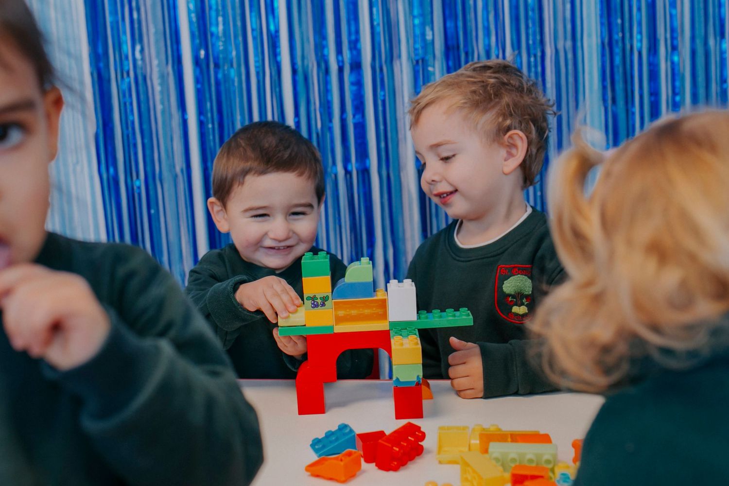 Young students in green uniforms play together with colorful building blocks at a classroom table.