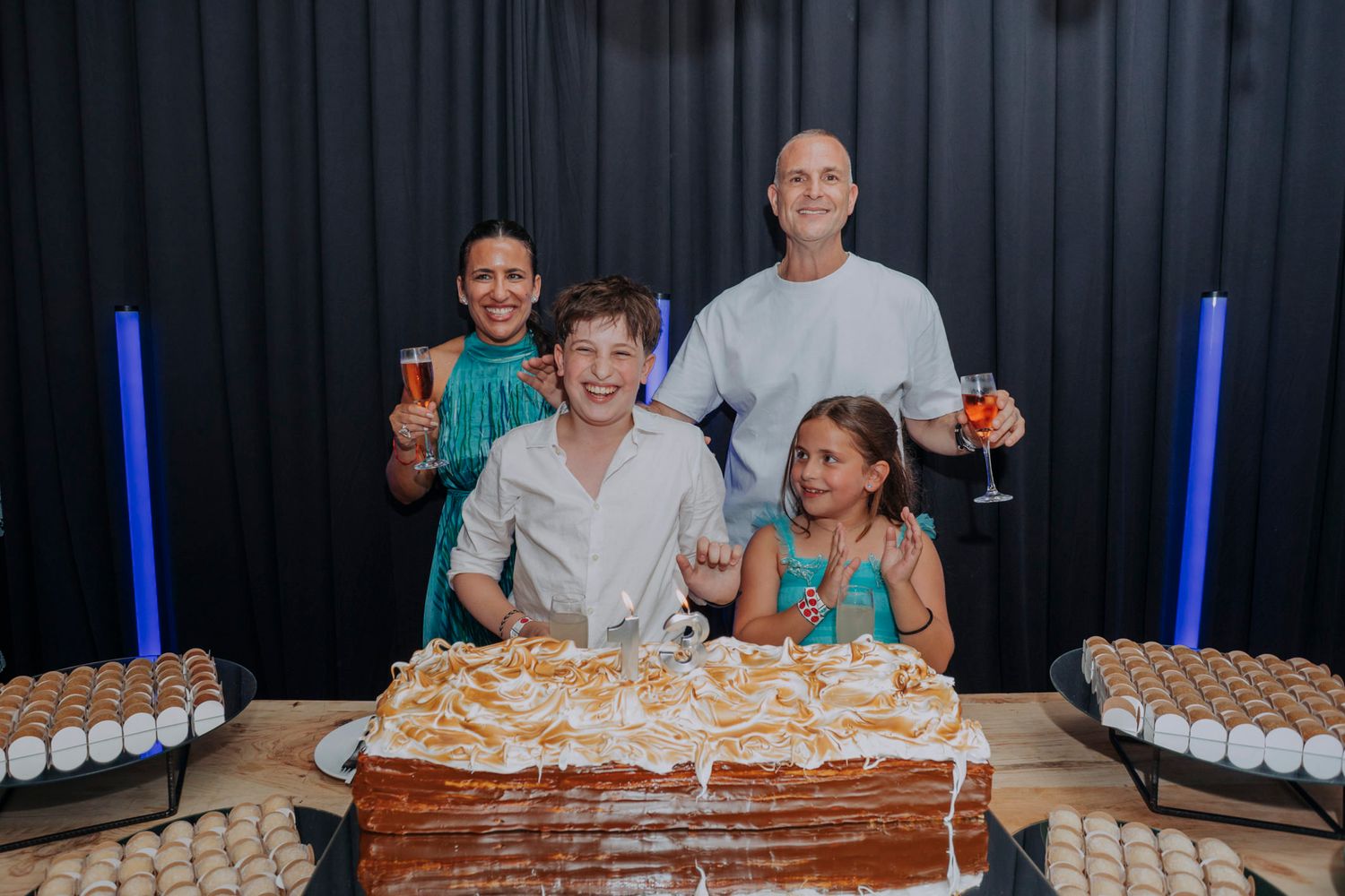 Family of four celebrating with a large layered cake, two adults holding cocktails and two children smiling at a festive party.