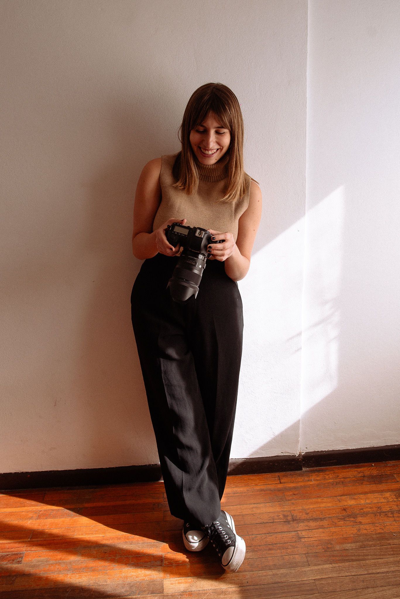 Professional photographer in dark pants smiling while checking camera settings in natural window light.