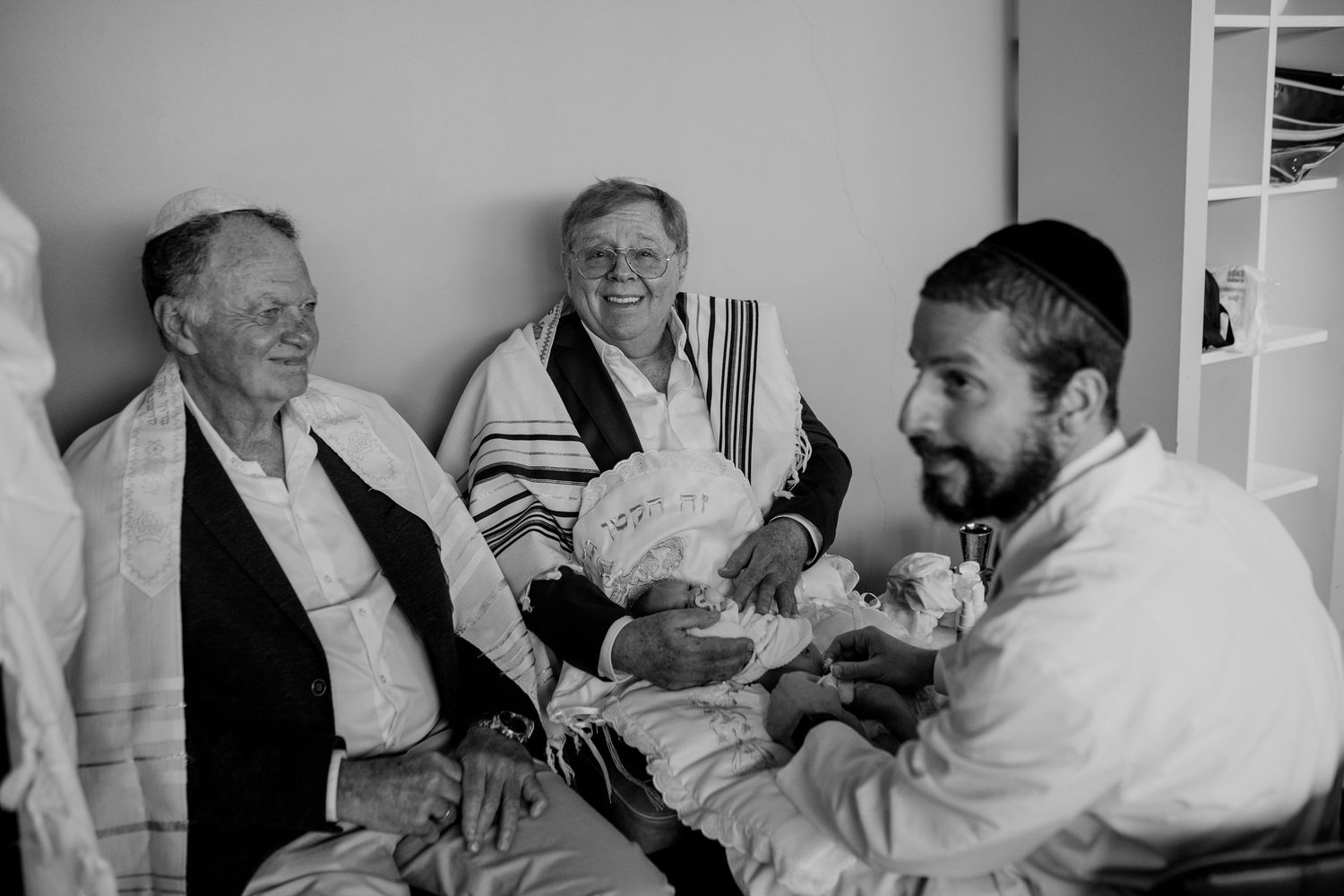 Black and white photo of men in Jewish prayer shawls celebrating a bris ceremony with a newborn baby.