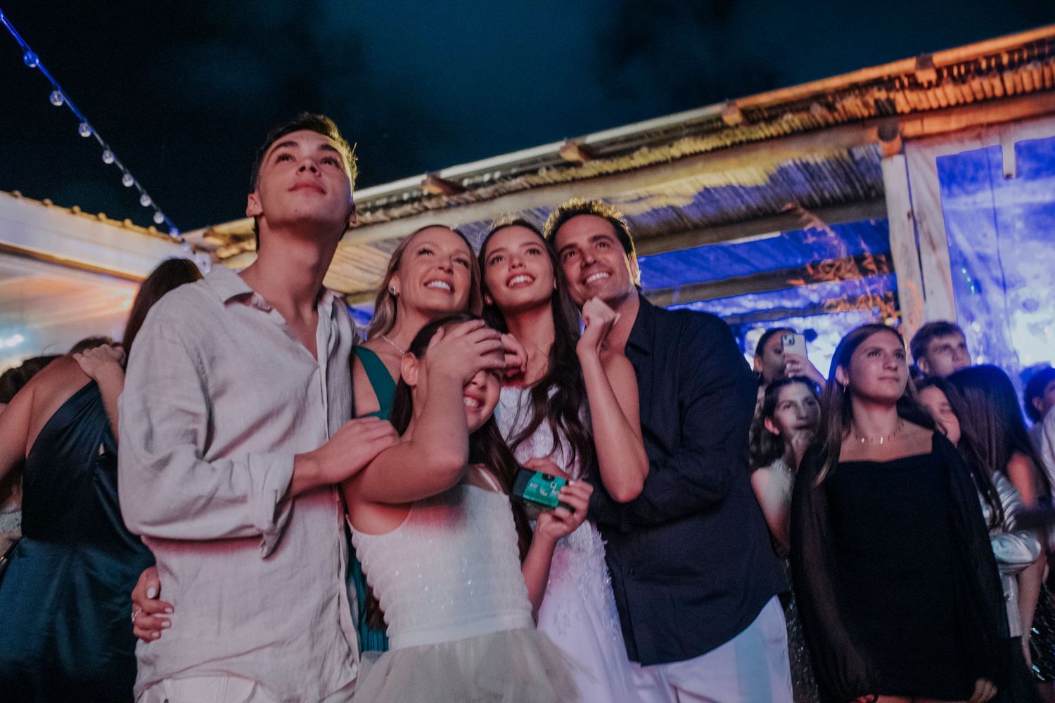 Party guests singing and celebrating together at a festive nighttime event with string lights overhead.
