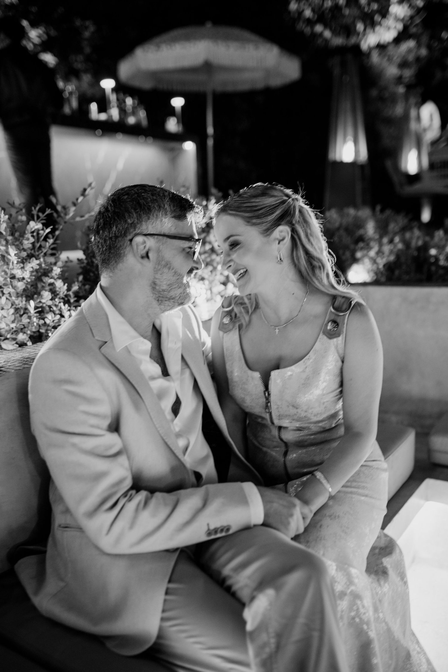 Black and white photo of a smiling couple in formal attire sitting closely together at an outdoor evening event.