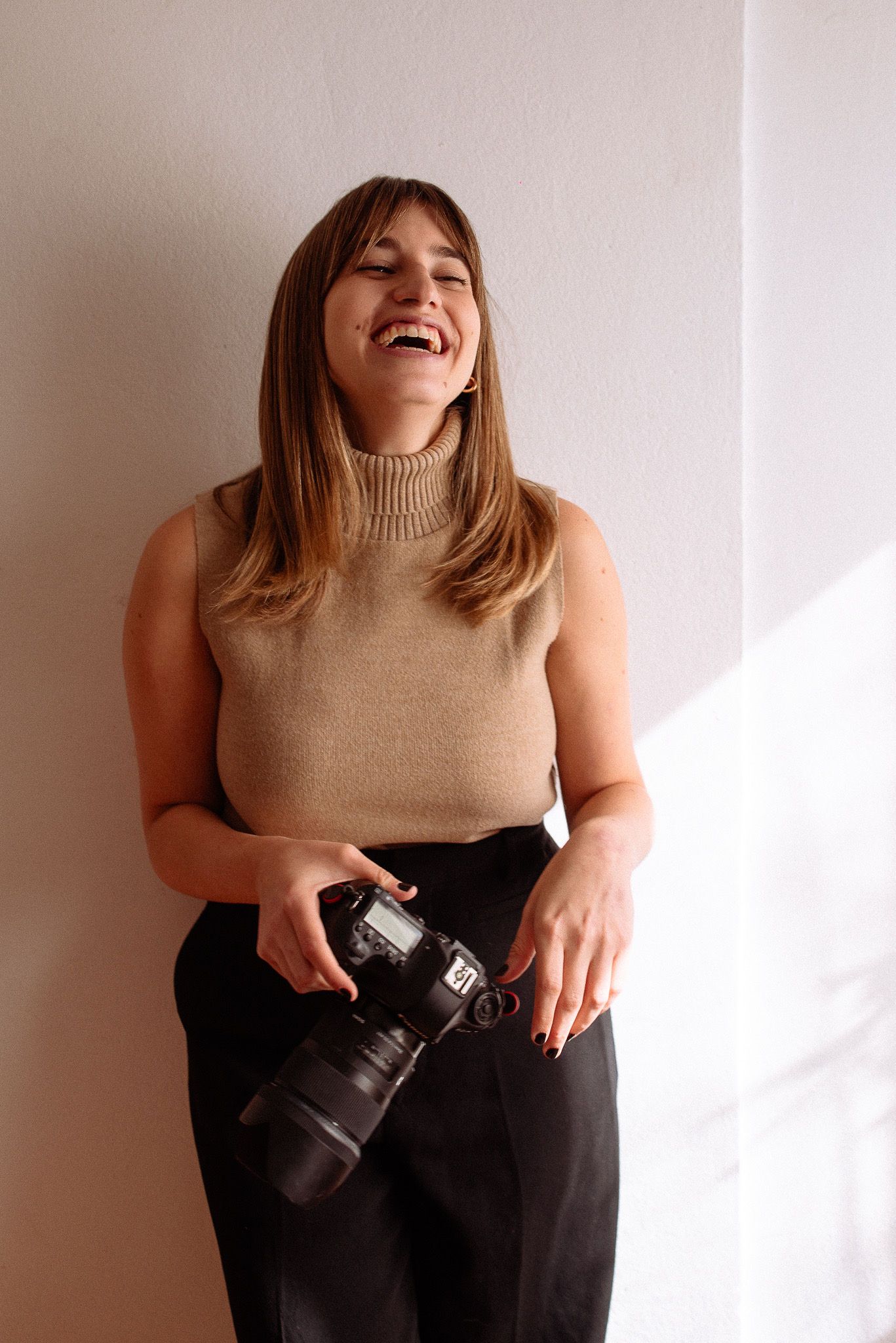 Professional photographer in a beige turtleneck holding a camera and laughing against a white wall.