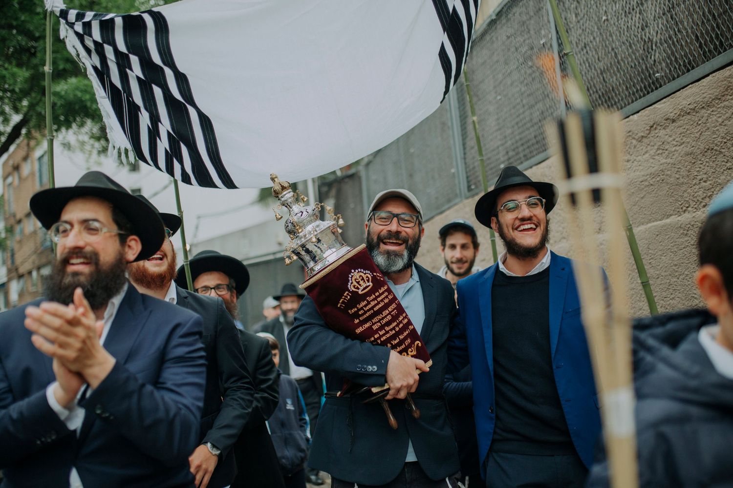 Jewish men celebrate outdoors, one holding a trophy, with a black and white flag waving above during a festive procession.