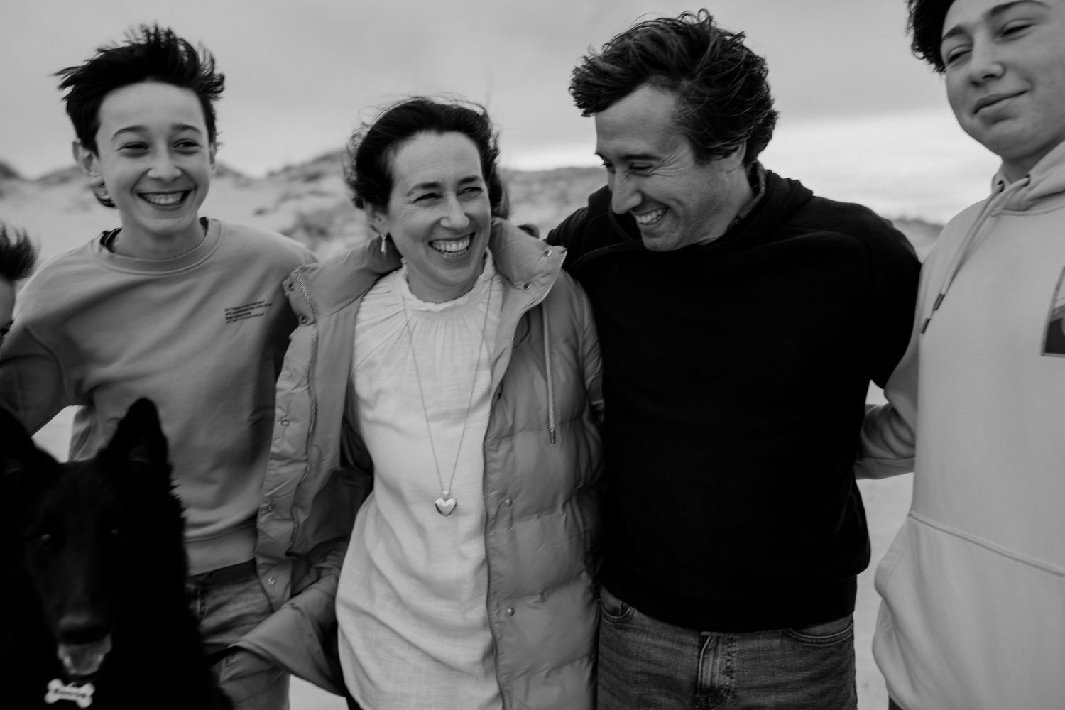 A black and white candid photo shows a joyful family laughing together outdoors on a bright day.
