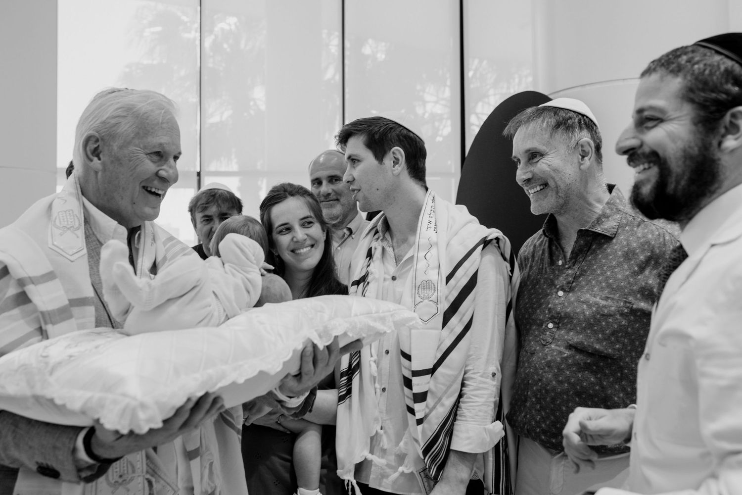 Jewish family celebrating a bris ceremony, with men wearing tallits, smiling and gathered around a newborn baby.