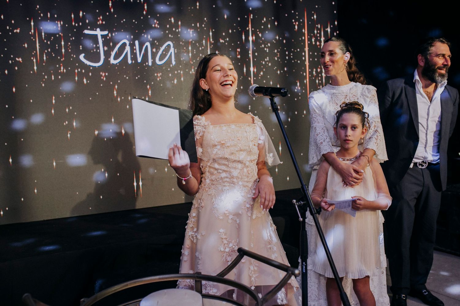 Young girl in white dress performs on stage at event with name Jana displayed on backdrop behind her.