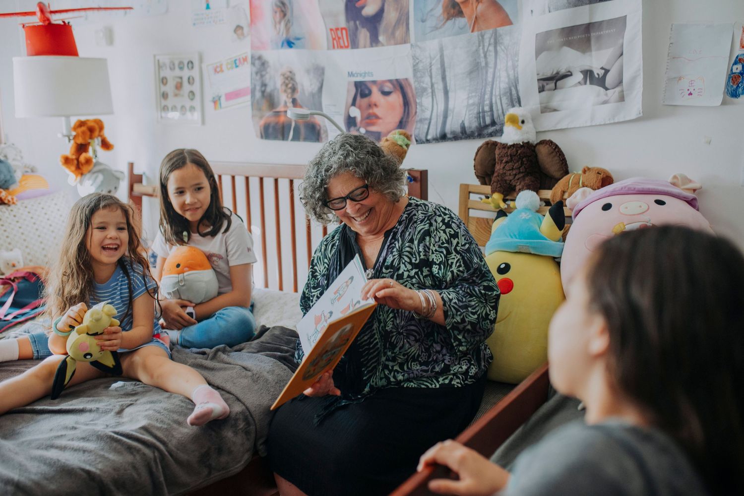 Grandmother reading a storybook to three young children surrounded by stuffed animals in a decorated bedroom.