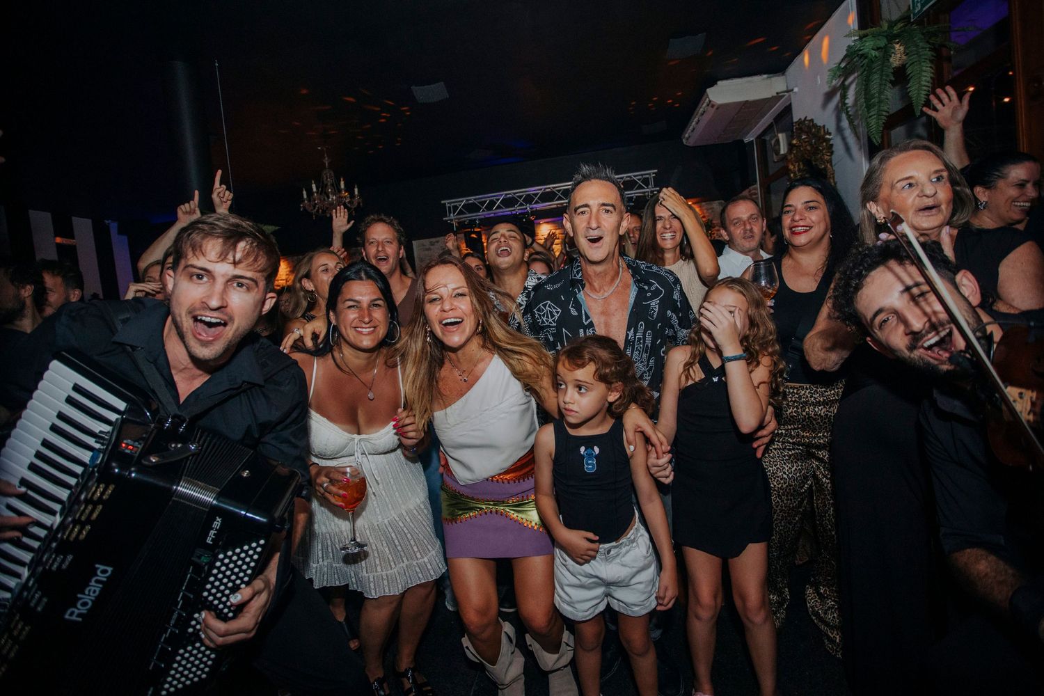 Lively party crowd with accordion player and violinist performing at an indoor venue with guests dancing and smiling.