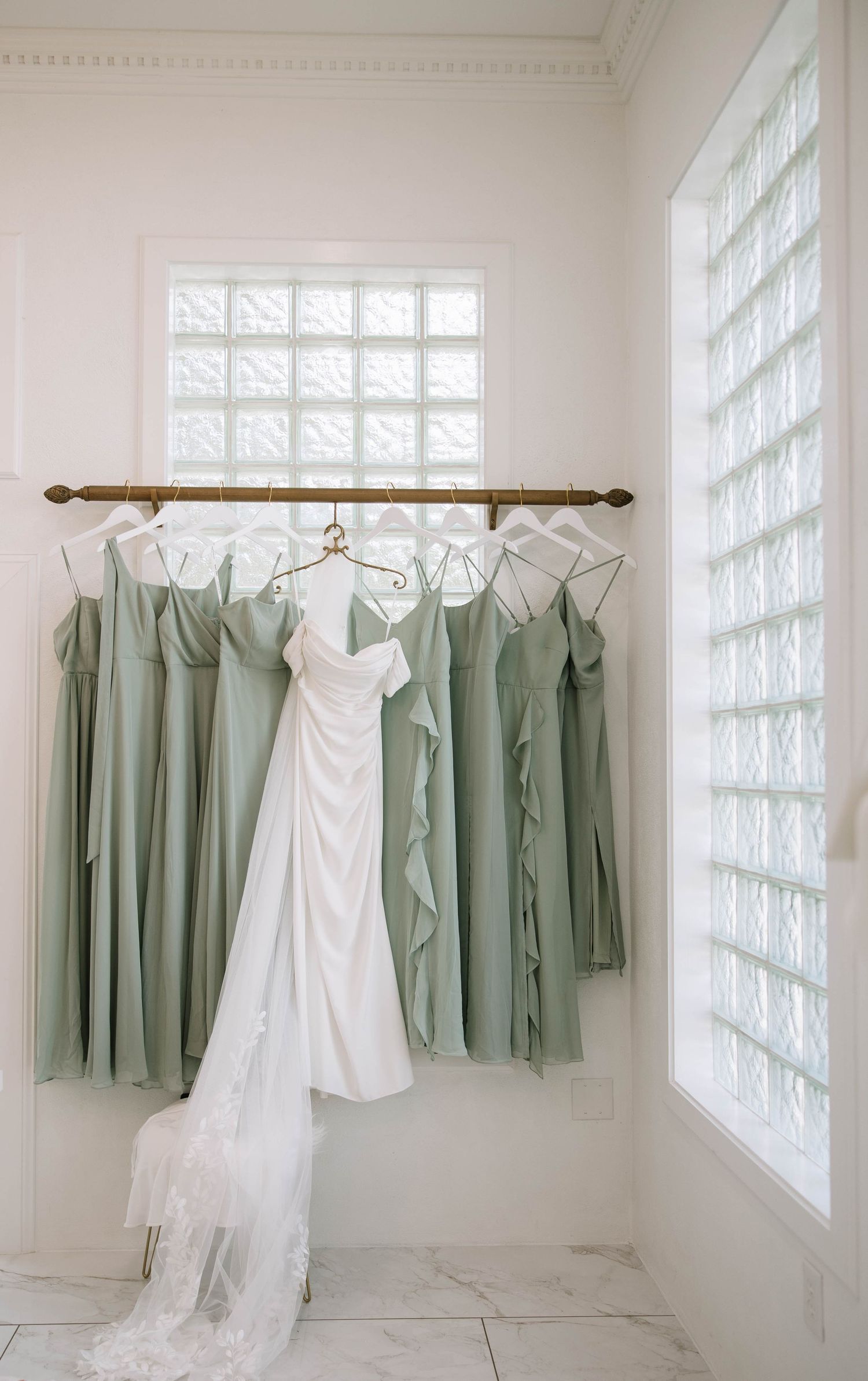 Wedding dresses and sage green bridesmaid gowns hanging on wooden rod in white room.