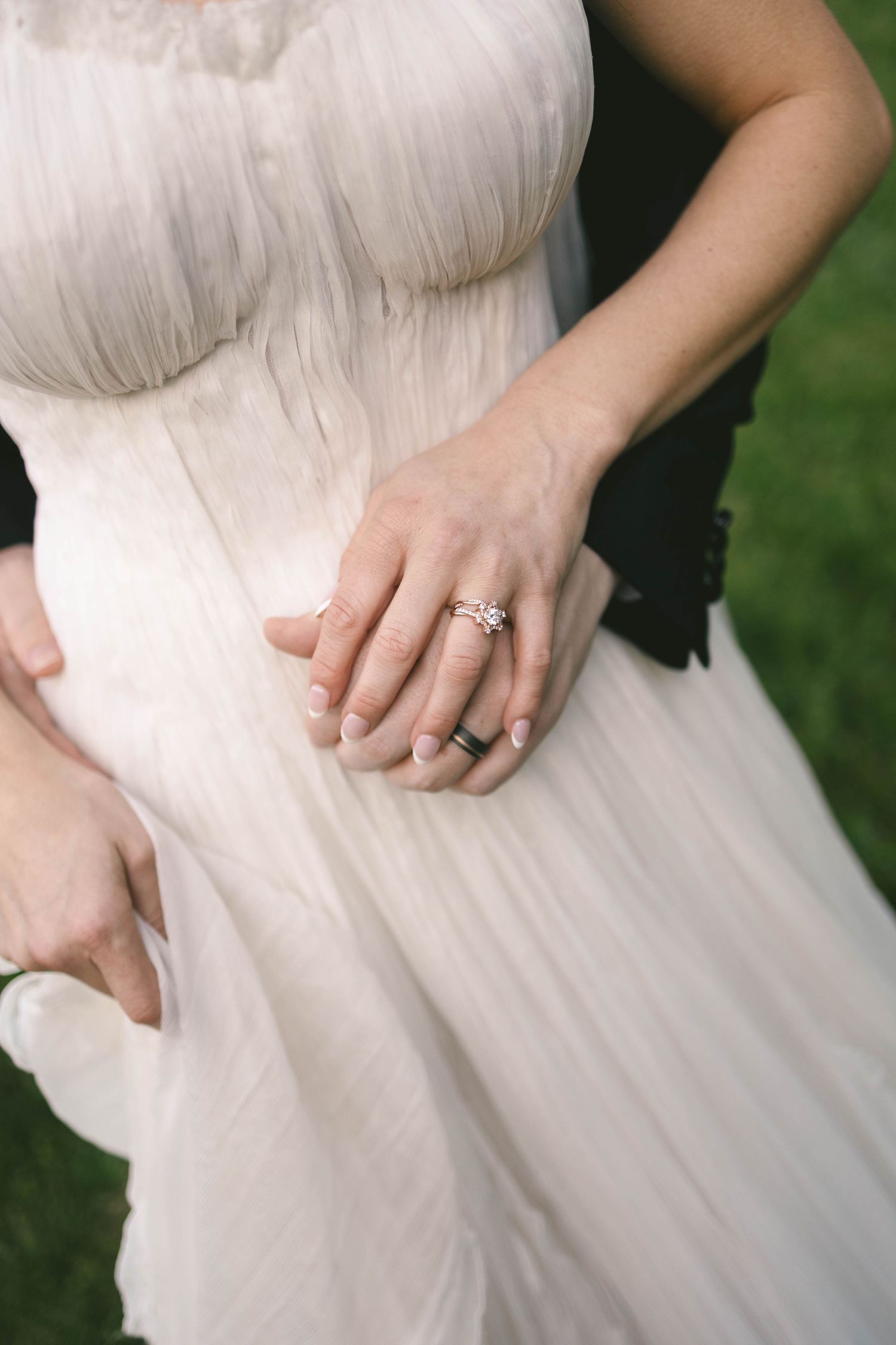 Close-up of bride and groom hands with elegant diamond engagement ring on white wedding gown.
