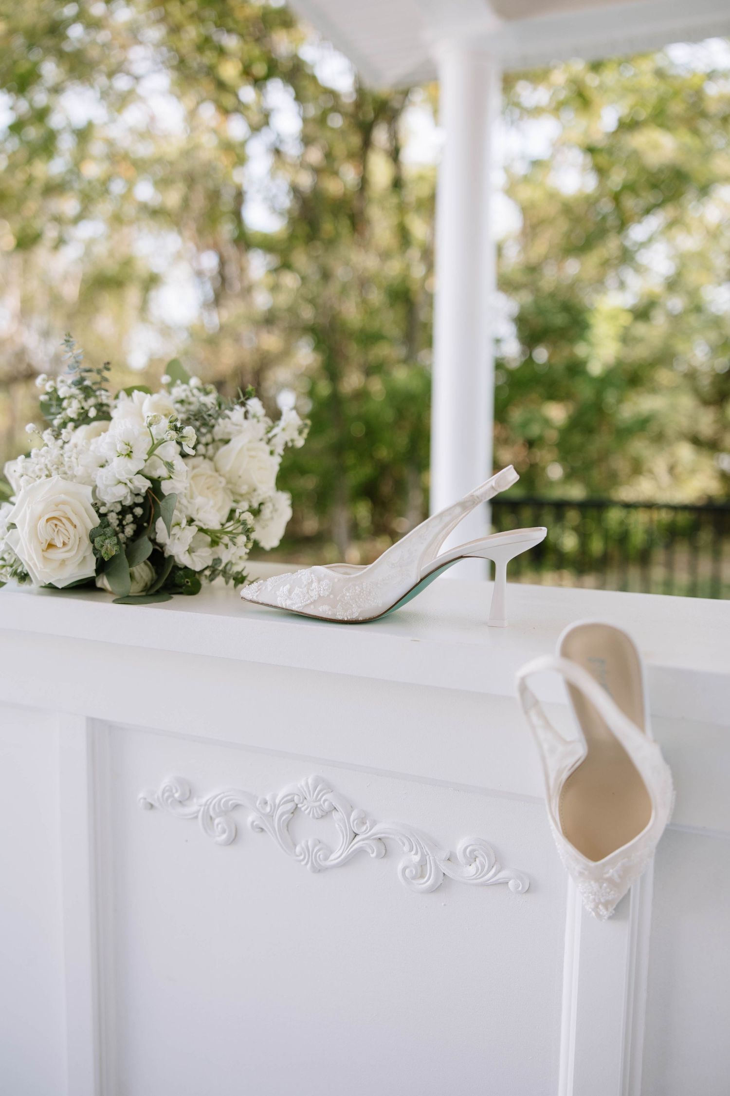 Elegant white bridal shoes and floral bouquet arranged on white table with decorative trim at outdoor wedding venue.
