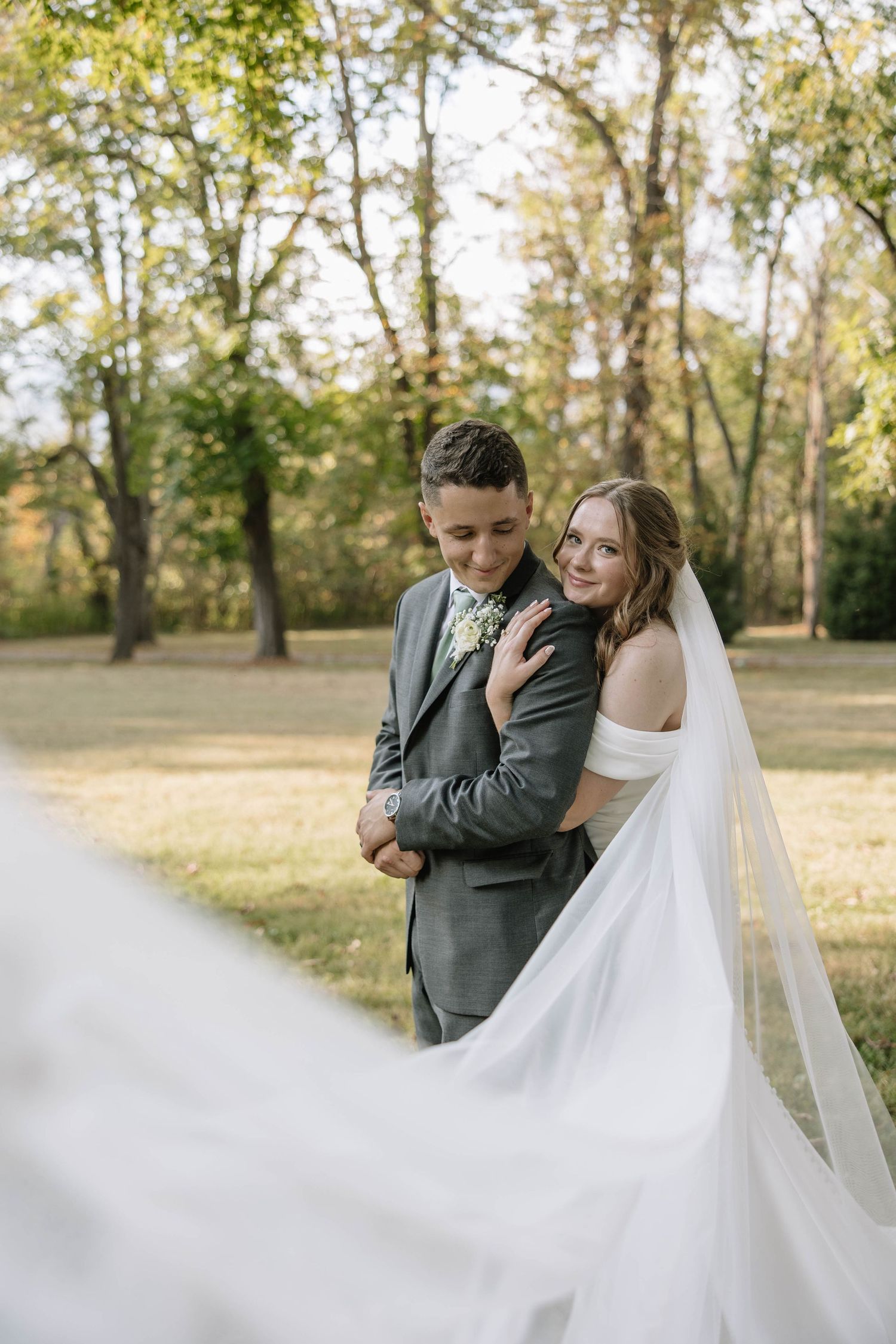 Wedding couple sharing tender moment with flowing white veil in natural outdoor setting.