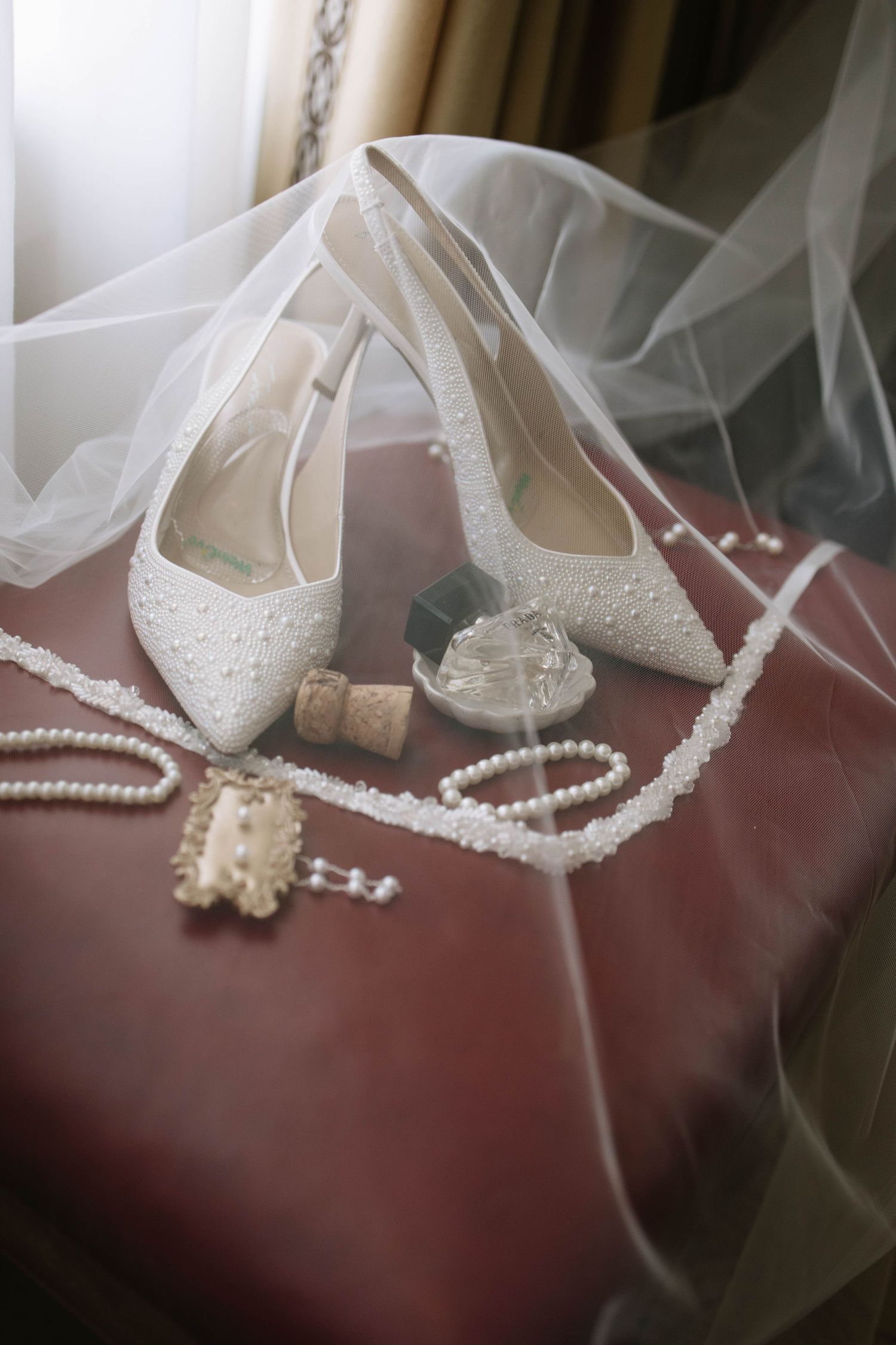 White lace bridal shoes and pearl jewelry arranged on red surface with delicate veil.