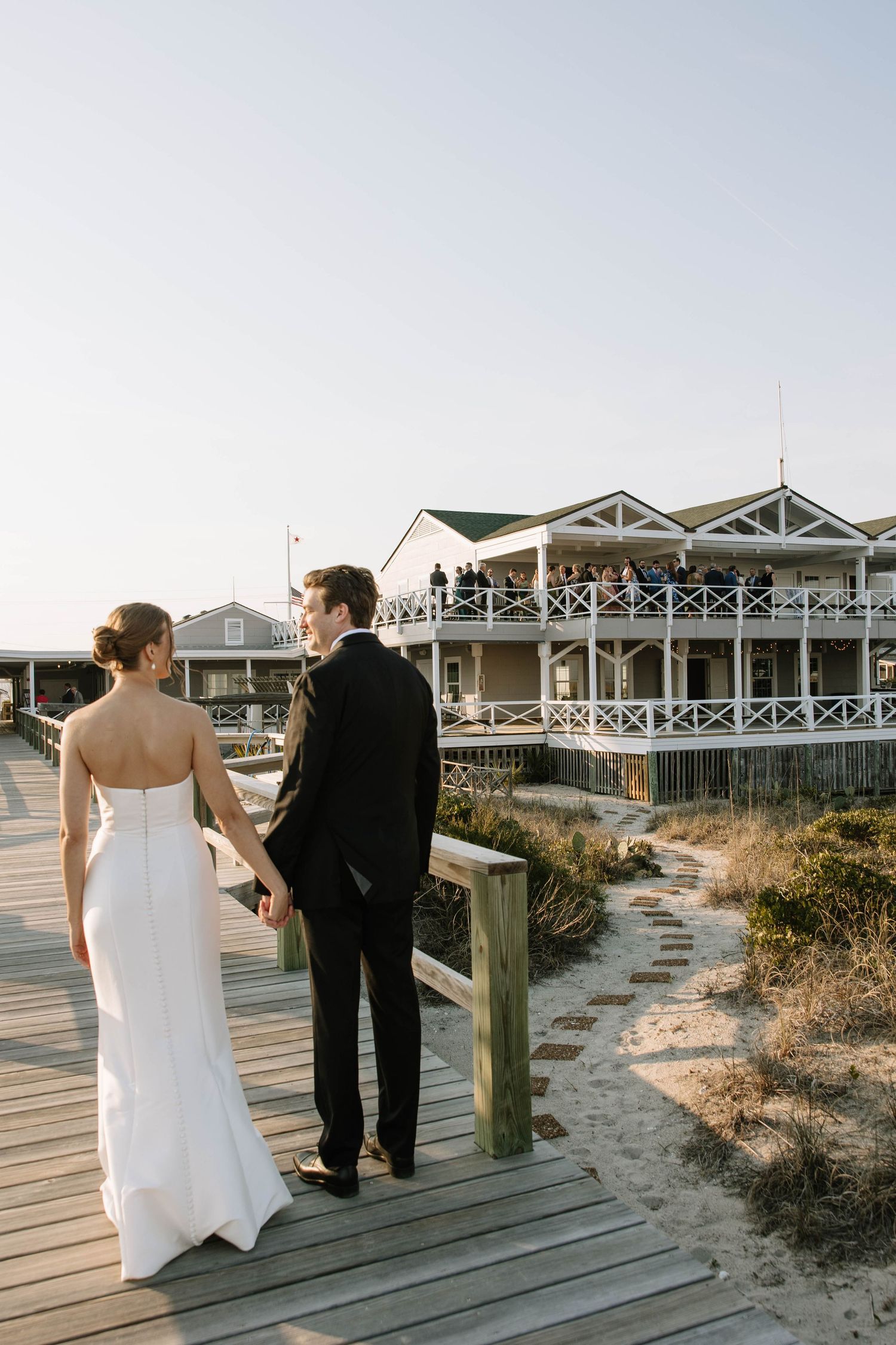 Bride and groom holding hands on a wooden boardwalk leading to a beachside venue with guests on the upper deck.
