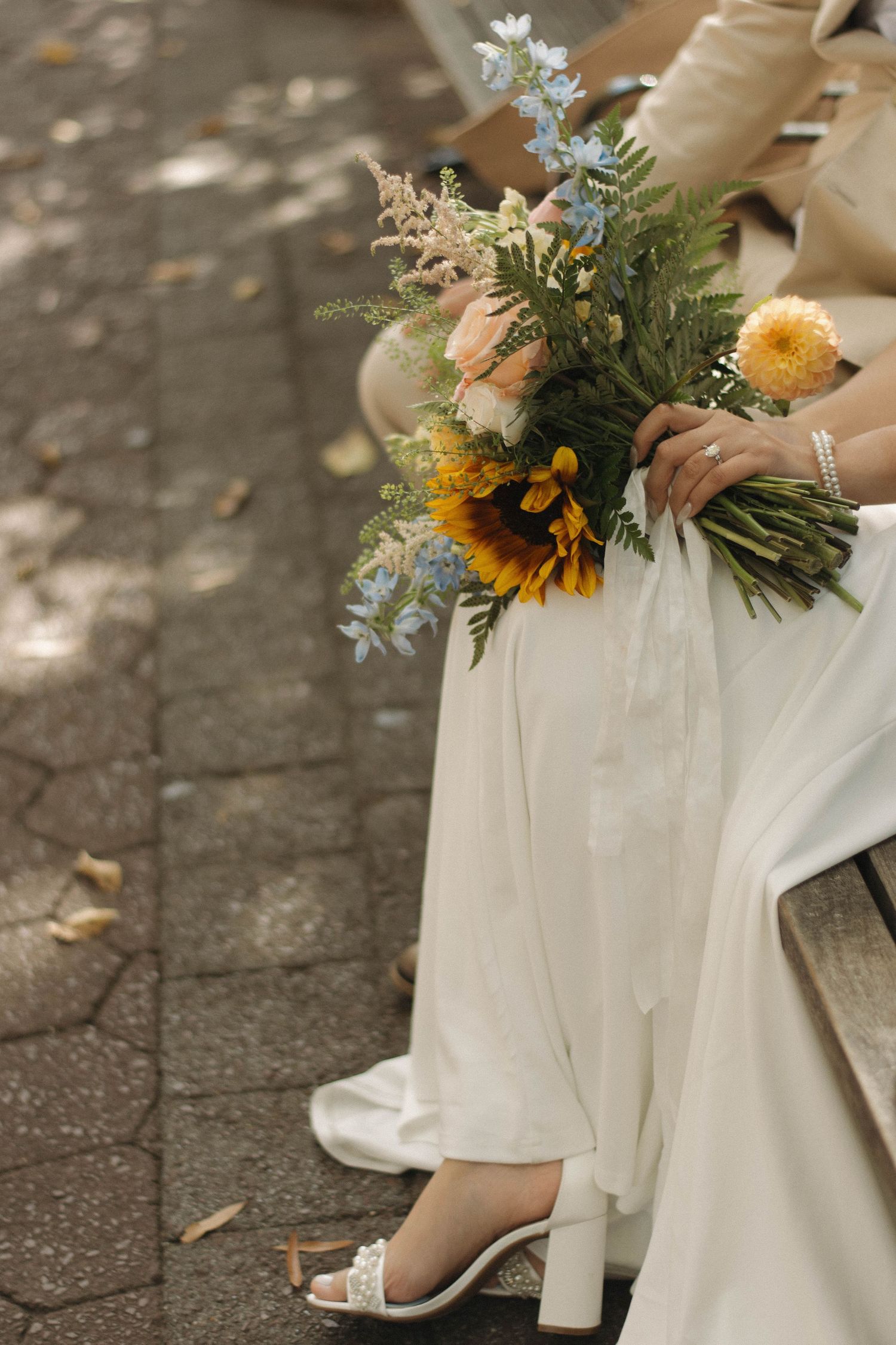 Elegant white wedding dress detail with sunflower bouquet and heeled shoes on stone pavement.