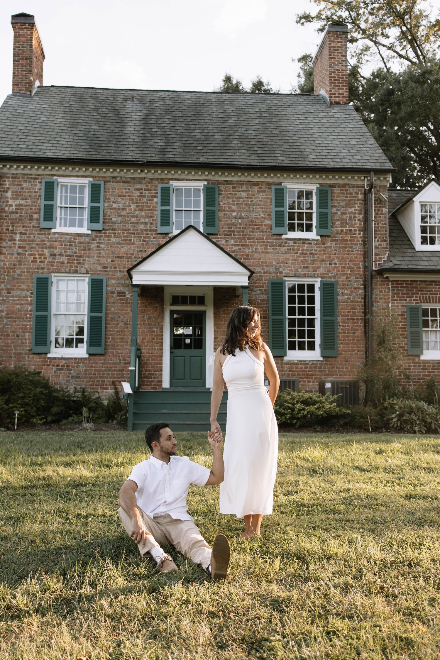 Couple poses in front of historic brick house with blue shutters during outdoor photoshoot.