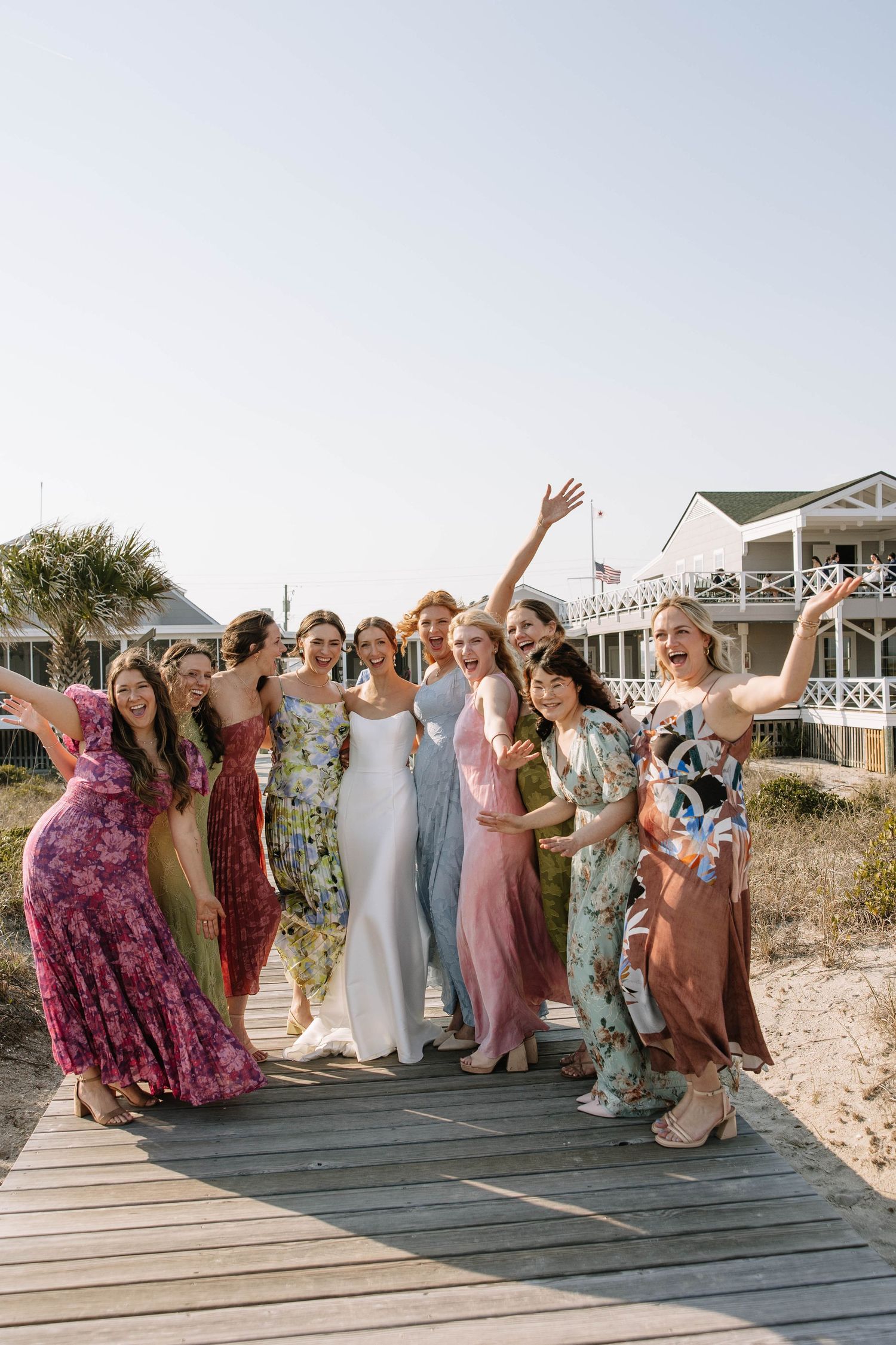 Bride and bridesmaids in colorful dresses celebrate on a beachside boardwalk on a sunny wedding day.