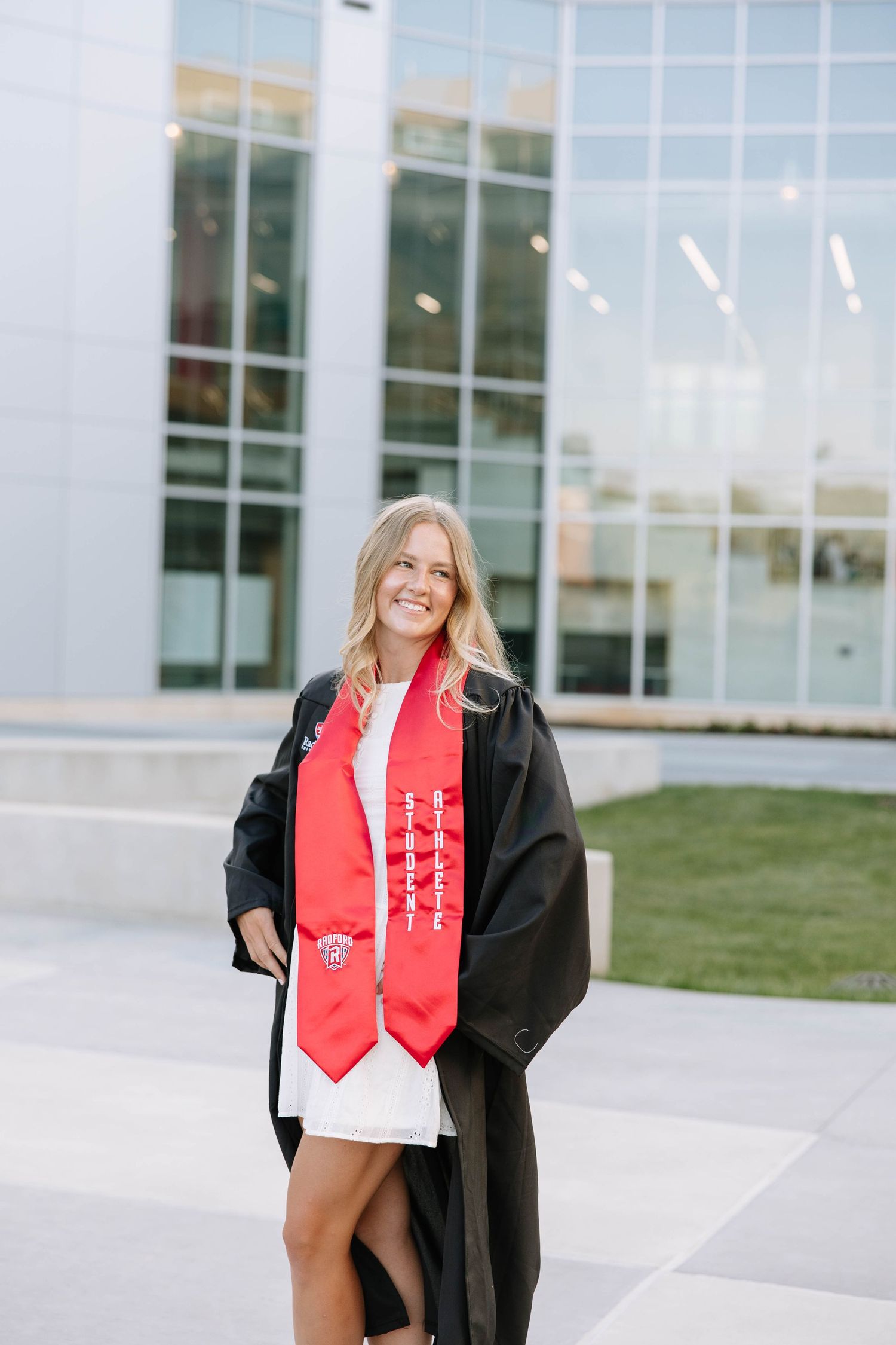 Smiling female graduate in black gown and red honor stole poses outdoors in white dress at university campus.